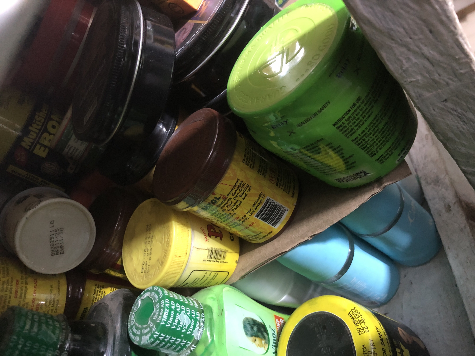 The image is a close-up, top-down view of a collection of cosmetic and medicinal products, primarily contained in various jars, bottles, and tubs. The products are densely packed, creating a cluttered composition. Visible items include a green plastic bottle with a label featuring a woman's profile and the text "Vinoz AD," several yellow tubs with labels that are partially obscured but appear to have product information and manufacturing dates, a larger green tub with embossed text including "SEALED FOR SAFETY" and "SO GOOD," and various dark-colored tubs with metallic rims. There are also blue cylindrical containers. Some of the labels are in English, and some appear to be in French. The items are arranged on a surface that appears to be made of cardboard or a similar material, suggesting they are stored in a box or on a makeshift shelf. The lighting is somewhat dim, casting shadows and creating highlights on the plastic and metallic surfaces. There are no people or animals visible in the image. The overall impression is one of a personal stash or a small retail display of personal care items. The context provided regarding Ado, Nigeria, suggests a domestic or small business environment.