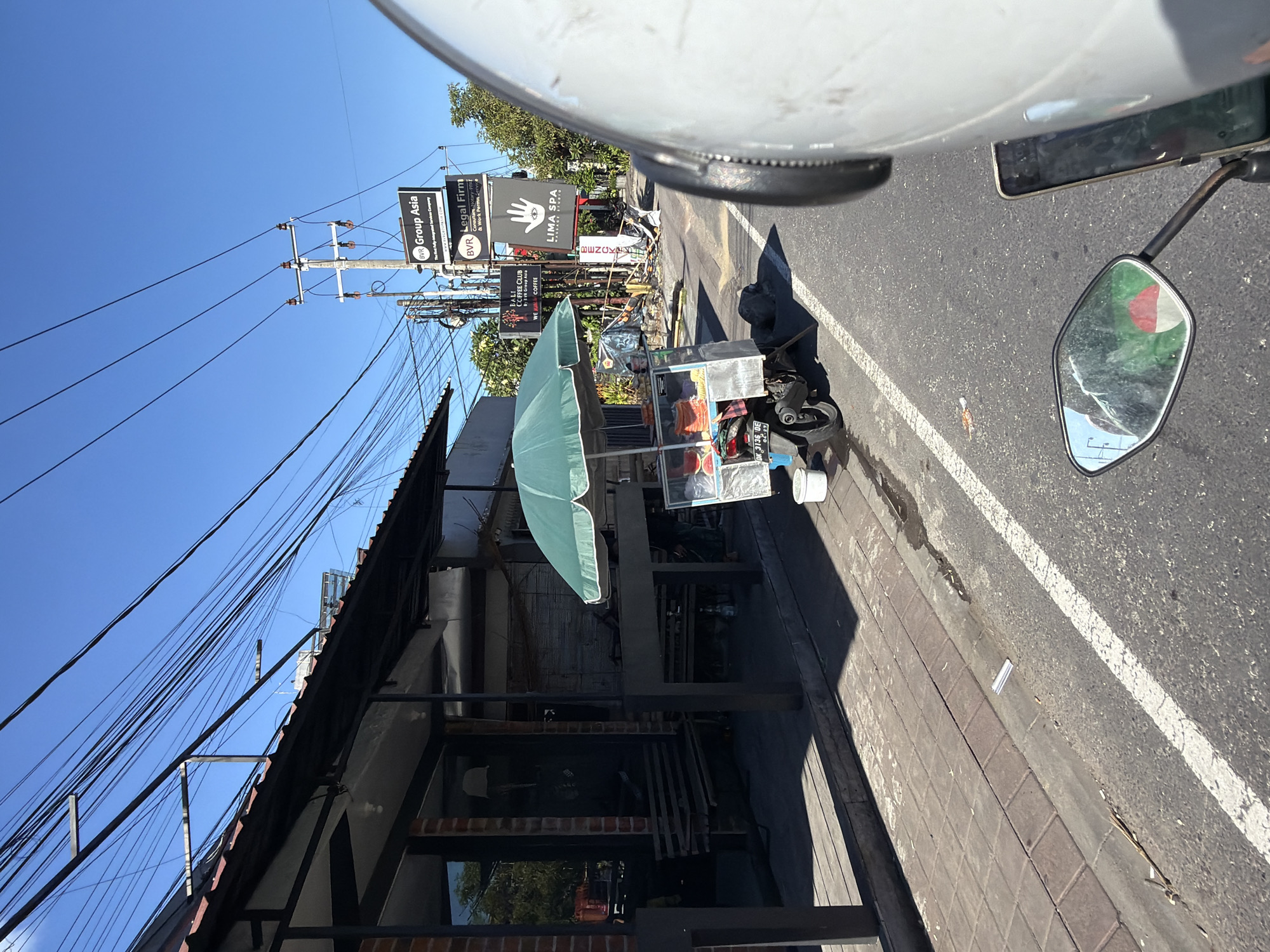 The image shows a street scene in North Kuta, Indonesia, viewed from the perspective of someone on a motorcycle.
Along the side of the road, there is a food cart shaded by a green umbrella. The cart appears to be selling fruits. Beside the cart is parked a scooter.
Various signs are visible, including "BVR Group Asia," "BVR Legal Firm," "LIMA SPA," and "BALI COFFEE CLUB."
Overhead are numerous power lines extending from a utility pole.
The road is marked with white lane lines.