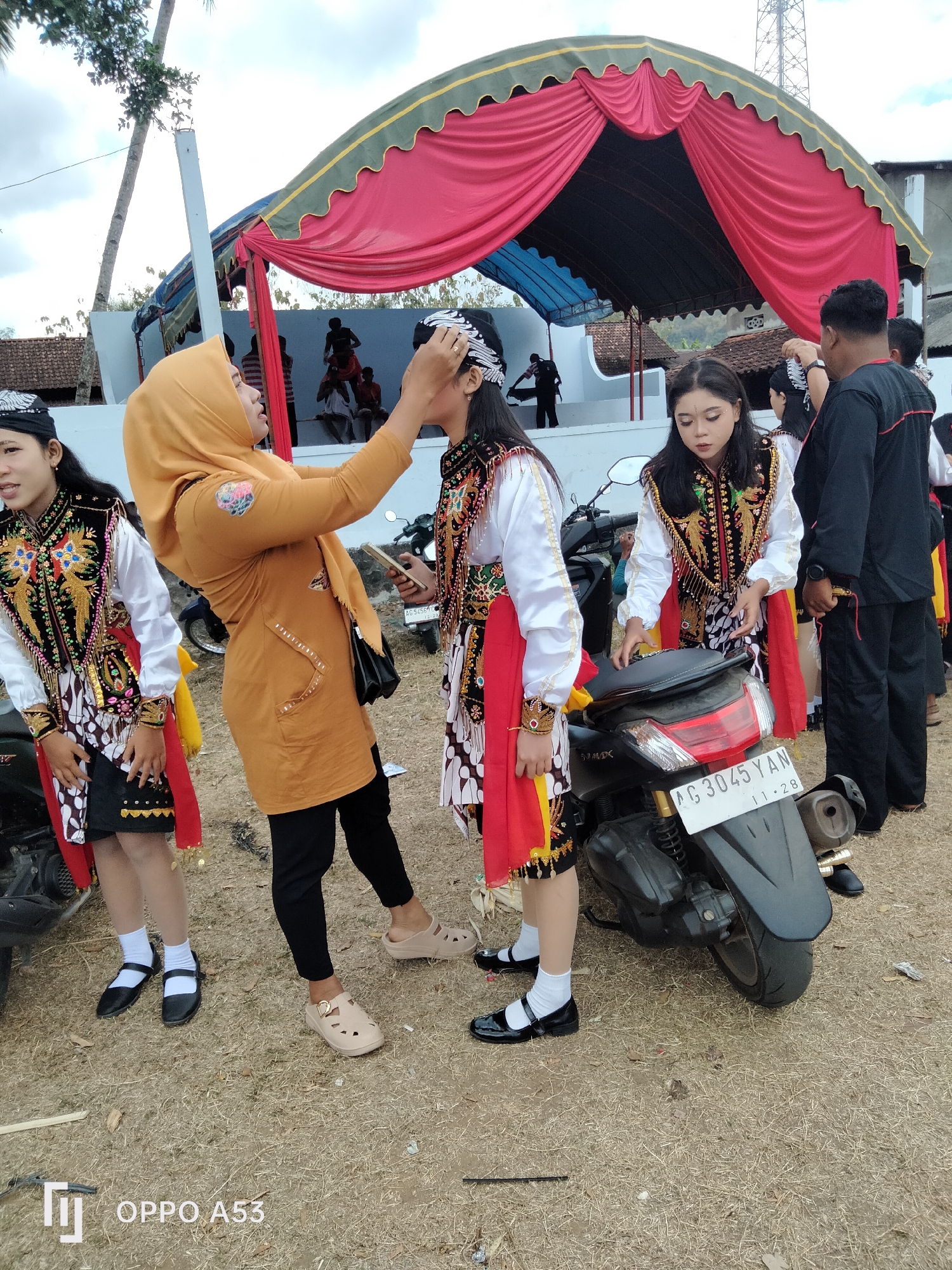 The image captures an outdoor scene in Bendoagung, Indonesia, likely during a cultural event. In the foreground, two young women dressed in traditional Javanese attire, consisting of embroidered vests, white shirts, and patterned skirts with red sashes, are standing next to a parked motorcycle. One of the women is adjusting the headwear of the other. A woman in a mustard-colored hijab and a brown jacket is also present, interacting with one of the performers. In the background, a stage is set up with a red and green draped canopy, and silhouettes of people are visible on it. The ground is covered with dry grass and scattered debris, suggesting a rustic or open-air setting. The overall atmosphere appears to be one of preparation or backstage activity before a performance.