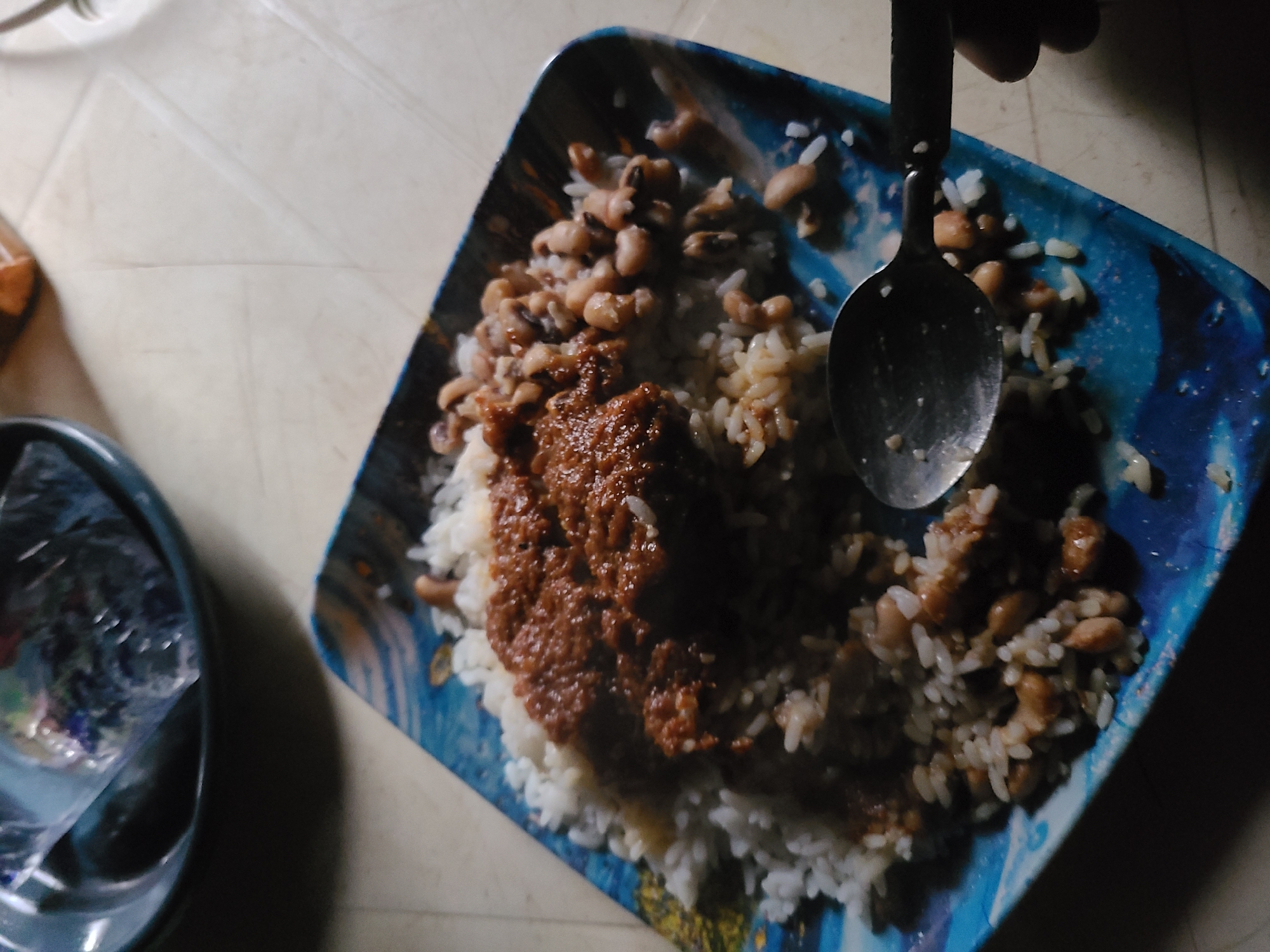 A top-down medium shot shows a square, blue-patterned melamine plate on a light-colored surface. The plate holds a serving of rice, topped with a dark-brown stew-like sauce and cooked black-eyed peas. A silver spoon rests on the food. To the left, a dark-colored bowl is partially visible, holding items obscured by plastic wrap. A hand is slightly visible on the right edge. The photograph may have been taken indoors. Specific location details within Jos, Nigeria, cannot be determined from the image.