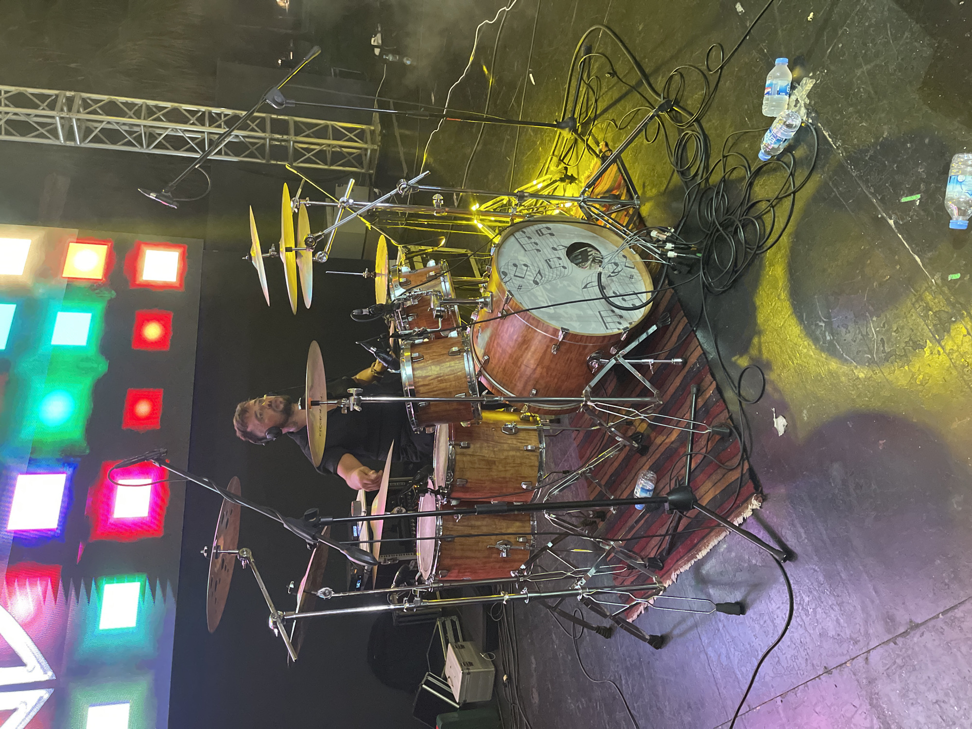 The image depicts a drummer performing on a stage in what appears to be an outdoor or semi-outdoor venue in Serik, Turkey. The drummer, wearing headphones, is seated behind a drum kit. The drum kit consists of several wooden drums with visible grain and metal cymbals mounted on stands. Microphones are positioned to capture the sound of the drums and cymbals.
In the background, a large screen displays colorful, abstract geometric patterns, primarily red, white, green, and pink squares. To the right of the screen, a metal truss structure is visible, suggesting stage lighting or rigging. The stage floor is dark and appears to be made of wood or a similar material, with various cables and wires strewn across it. Yellow and purple stage lights cast shadows and illuminate the scene.
In the foreground, on the stage floor, are several empty plastic water bottles and coiled black cables. The overall atmosphere is that of a live music performance.