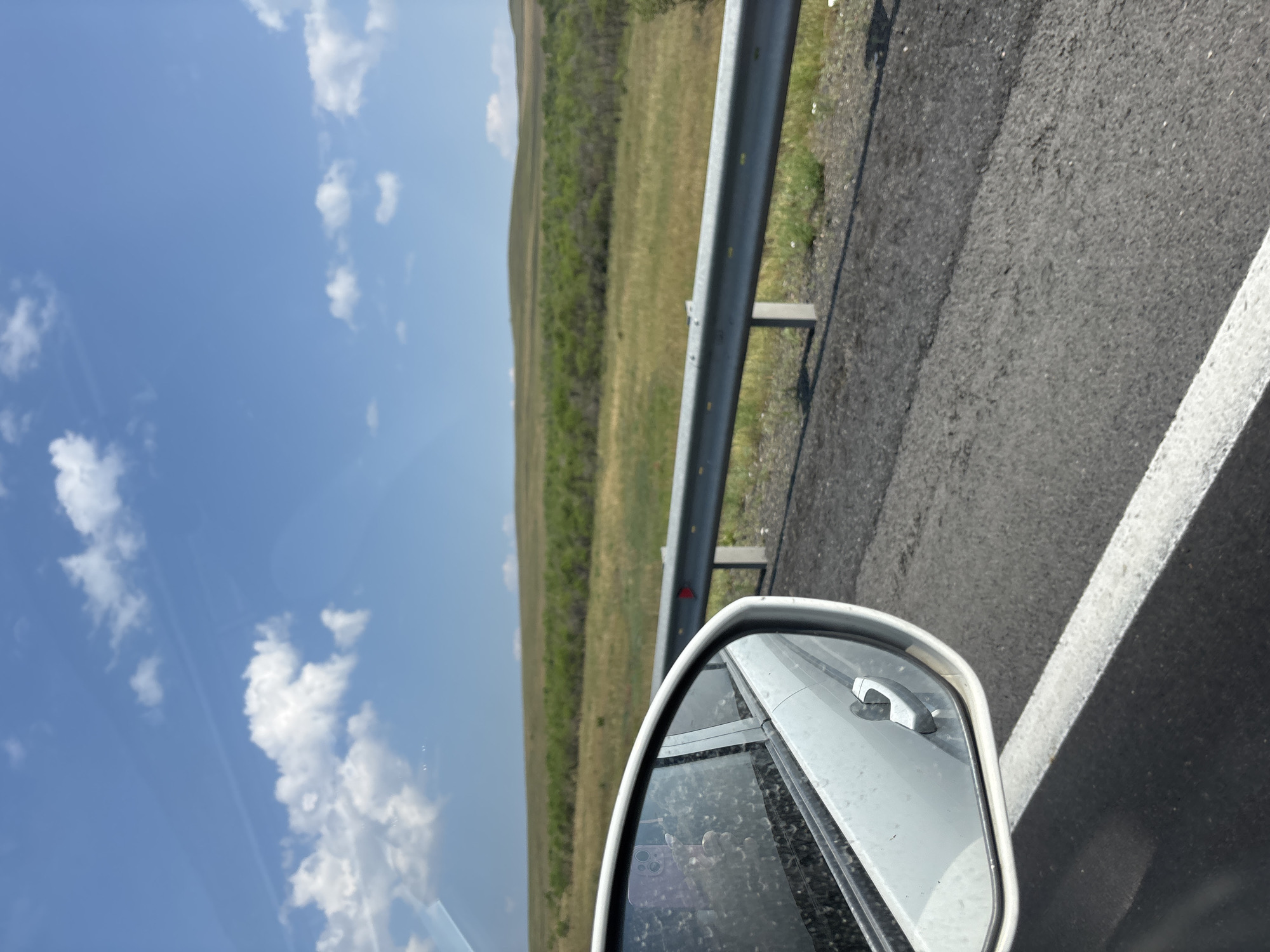 The image is a forward-facing view from a vehicle, looking out at a landscape. In the foreground, the side mirror of a white car is visible on the left, reflecting a hand holding a purple phone. The mirror is speckled with water droplets, suggesting recent rain or washing. The car's door handle is also visible. The road surface is asphalt with a white lane marking. A silver metal guardrail runs along the right side of the road. Beyond the guardrail, the landscape consists of rolling green hills covered in sparse vegetation, with a line of trees in the middle distance. The sky is blue with scattered white cumulus clouds. The scene is likely a roadside view in or near Chernogorsk, Russia, given the provided context of the city.