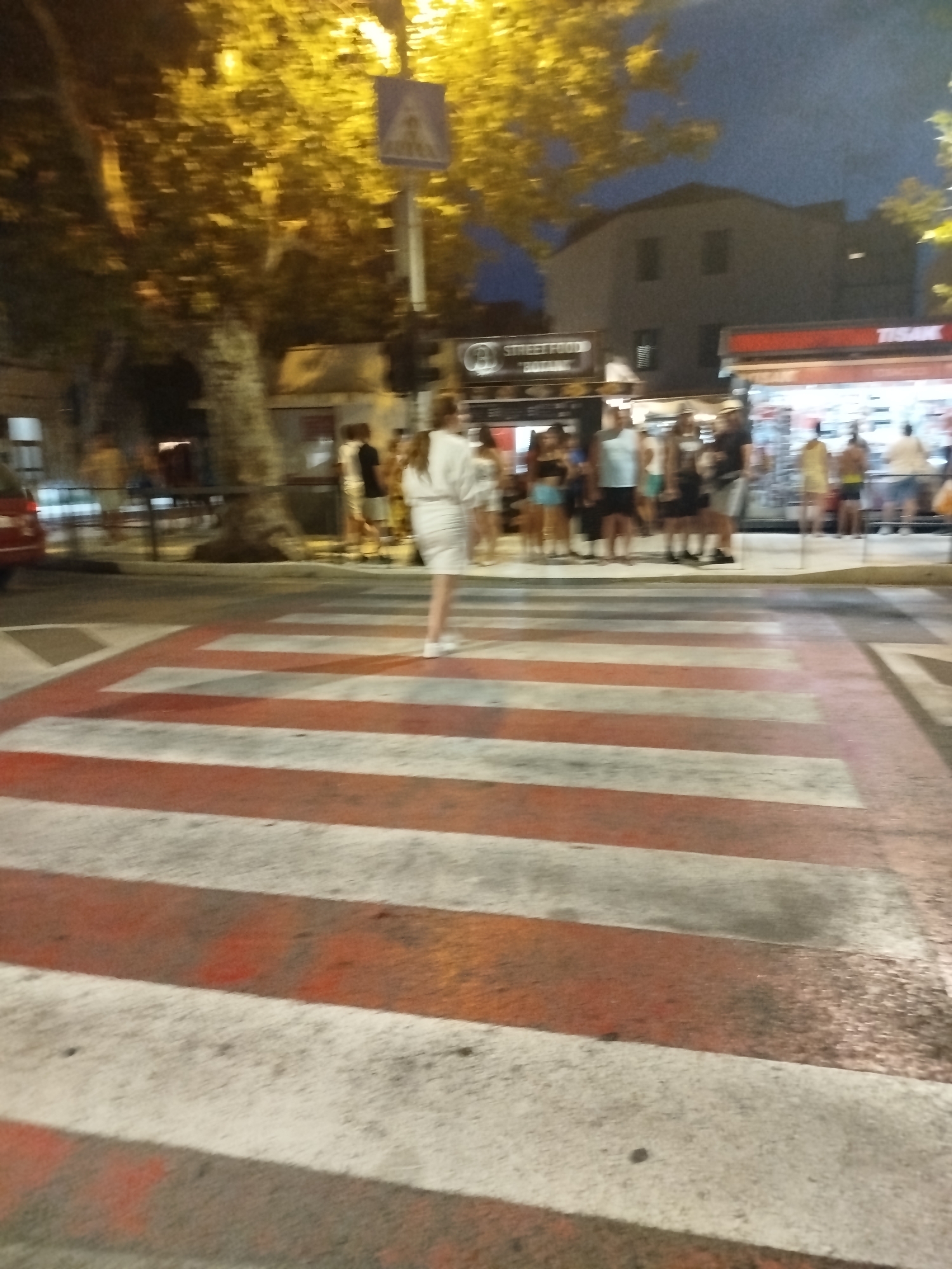 In Omiš, Croatia, at night, a crosswalk with alternating red and white stripes is illuminated by warm yellow streetlights. A blurry pedestrian in a white dress stands on the crosswalk, facing right. In the background, a group of people are gathered near food stalls with glowing signs, including one labeled "STREET FOOD." A red car is parked to the left of the frame.