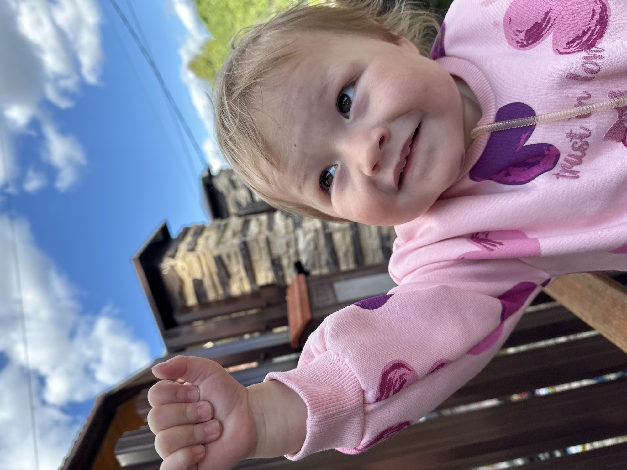 A smiling toddler with light hair and dark eyes is captured in a close-up, low-angle photograph taken outdoors in Nizhny Novgorod, Russia. The child looks directly at the camera with a cheerful expression, wearing a pink long-sleeved top or hoodie. This top is decorated with purple heart-shaped motifs, and the partially visible text "trust" and potentially "love" can be seen on the fabric, along with a transparent plastic drawstring near the neck.
The child's right hand is extended towards the camera, with fingers slightly curled, suggesting an interaction or playful gesture. They appear to be positioned near or on a dark wooden structure, possibly a bench or railing, which runs diagonally across the lower part of the frame.
In the background, under a clear blue sky filled with white, fluffy clouds, stands a prominent building. This structure features several levels and is constructed from textured, stone-like materials, giving it a rustic or historical appearance. Some green foliage is also visible in the upper part of the background. The scene is brightly lit, indicating a sunny day.
