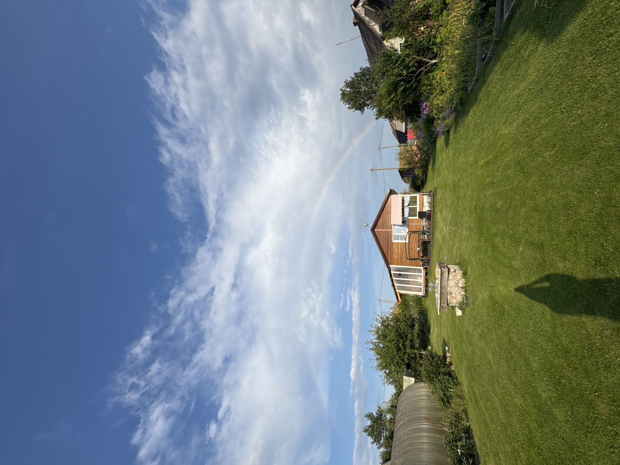 The image captures a rural scene in Sitno, Russia, with a vibrant rainbow arcing across a blue sky dotted with white clouds. In the foreground, a meticulously mowed green lawn leads up to a modest wooden house with a sunroom. To the left, a greenhouse or polytunnel stands partially visible. The house features a pinkish-brown exterior with white trim and windows. A stone well with glass jars sits on the lawn. In the background, other houses and trees are visible, contributing to the bucolic atmosphere. The shadow of a person holding a phone, presumably the photographer, is cast on the lawn. The overall impression is one of a peaceful summer day in the Russian countryside.