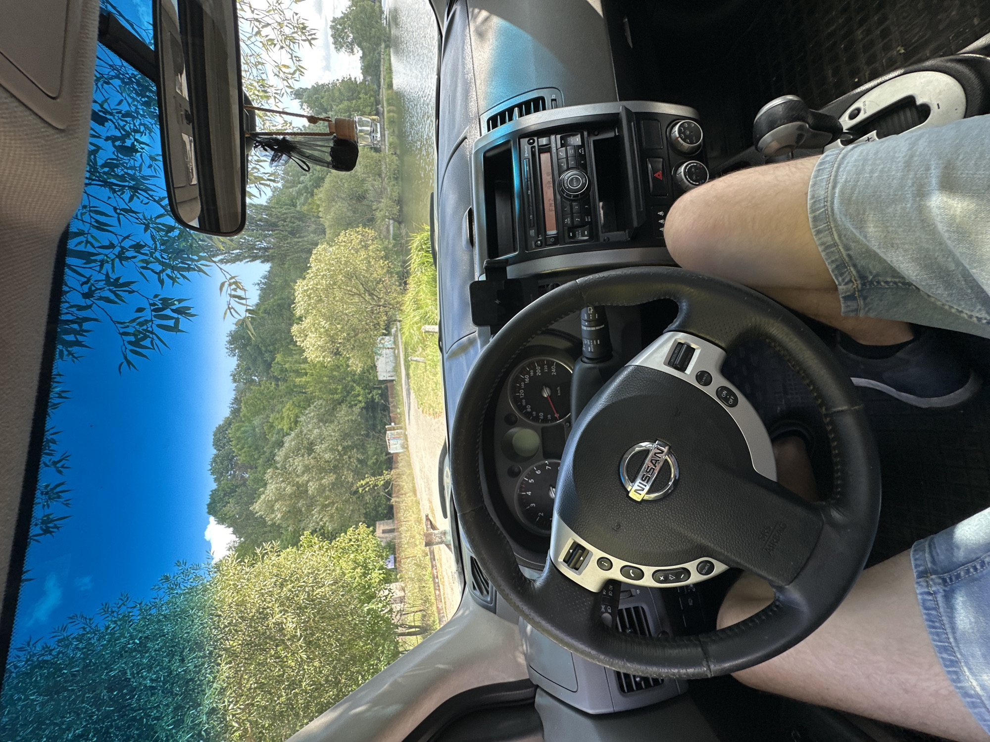 The image provides a first-person perspective from the driver's seat of a car, looking forward through the windshield. The vehicle's dashboard, steering wheel with the Nissan logo, and gear shift are prominently featured. The driver's legs, clad in denim shorts, are visible. Outside the car, a body of water, likely a lake or river, is visible, bordered by lush green trees and vegetation. The sky is a clear, bright blue with a few wispy clouds. The scene appears to be set in a natural, rural environment. There are no other people visible in the image. The location is identified as Makiv, Ukraine.