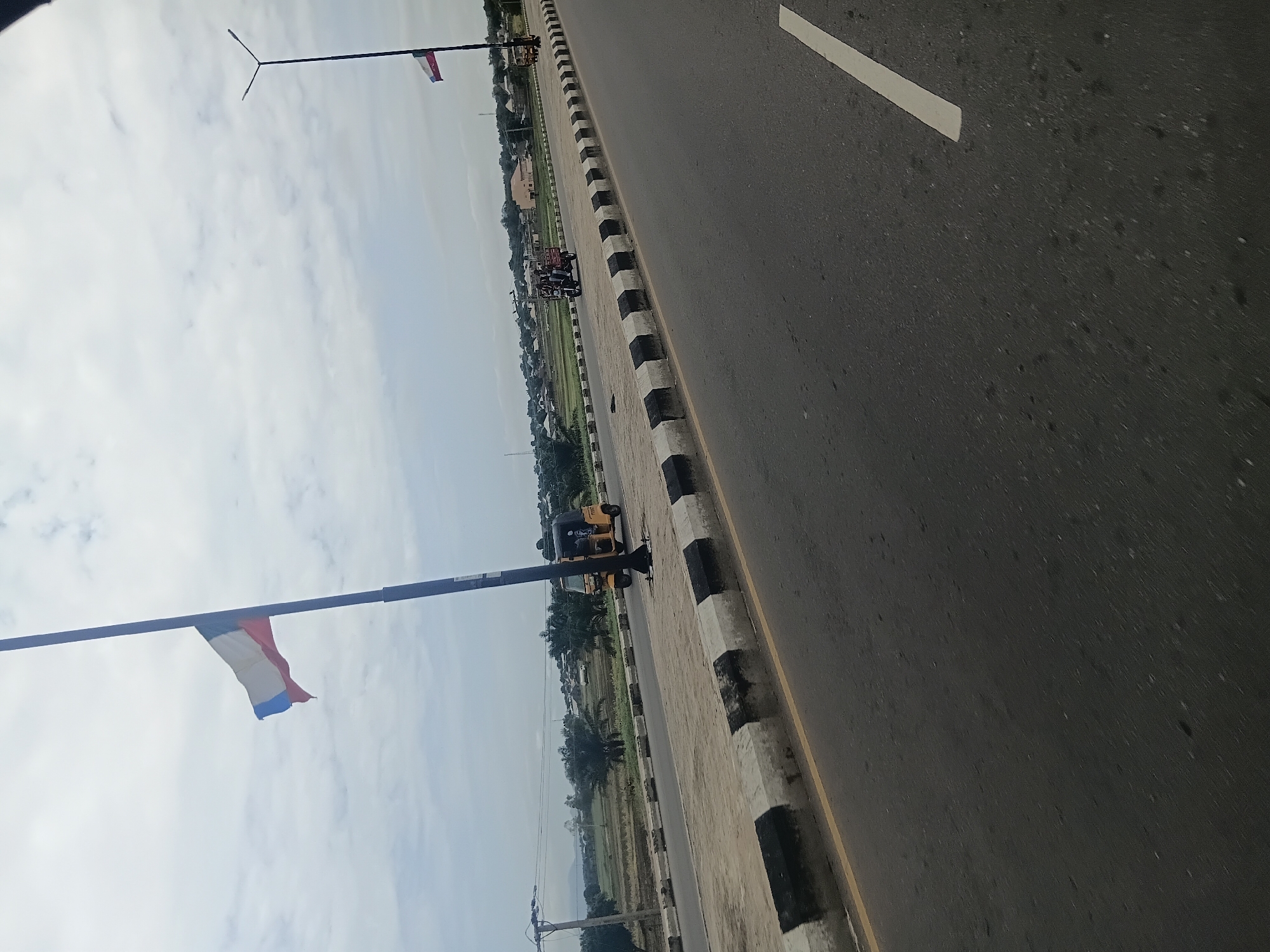 The image depicts a scene along a road in Jalingo, Nigeria. The perspective is from a moving vehicle, looking towards the left side of the road and into the distance.
The primary subjects include a paved road with a white line marking its edge. Adjacent to the road is a sidewalk with a distinctive black and white striped curb. Beyond the sidewalk, there are signs of a rural or semi-urban environment with scattered buildings, palm trees, and green fields, suggesting agricultural land. A light pole with a flag that appears to be red, white, and blue is visible in the foreground, attached to a dark pole. Further down the road, another light pole is present, and in the middle distance, several people are riding on a motorized tricycle, a common mode of transportation in Nigeria. A yellow tricycle is also parked near the sidewalk.
The activity appears to be daily life and travel on a road. The time of day is likely daytime, given the brightness and visible shadows. The sky is partly cloudy, indicating moderate weather conditions. The presence of local vehicles and vegetation points to a tropical or sub-tropical location. There is no visible text in the image. The overall impression is one of a typical day in a Nigerian town or its outskirts, with everyday activities and transportation taking place.