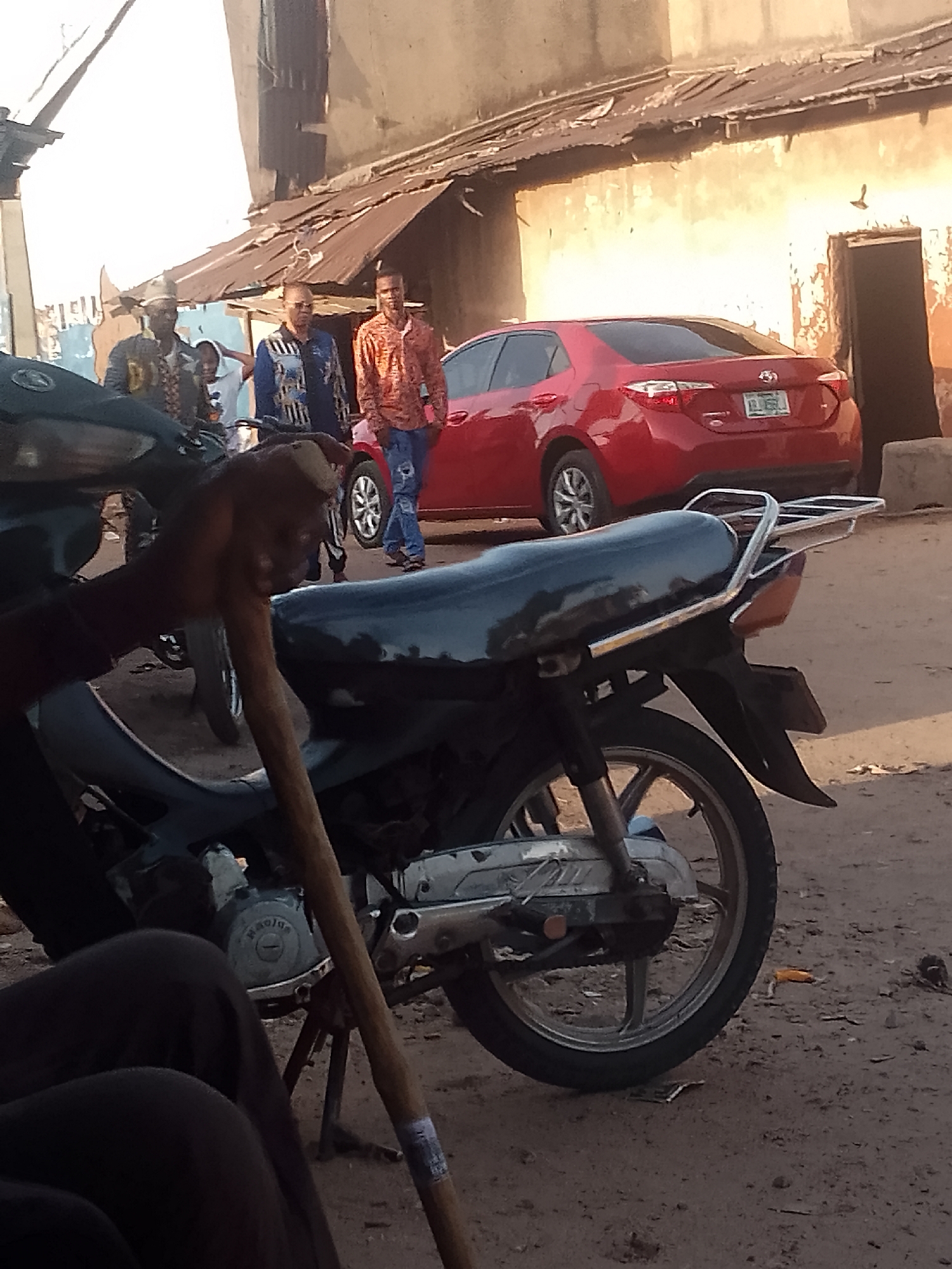 The image presents a street scene in an unknown city and country. In the foreground, a black motorcycle is parked, obscuring some of the view. A wooden walking stick rests against it. Behind the motorcycle, a red Toyota Corolla is parked near a weathered building with a door opening to the side. Several individuals are walking or standing near the car. One man wears a red patterned shirt with blue jeans, and another is in a blue and white patterned jacket. There appear to be two or three other individuals standing together on the left side of the frame, but they are more obscured. The road appears unpaved. The environment has a somewhat worn, urban feel.