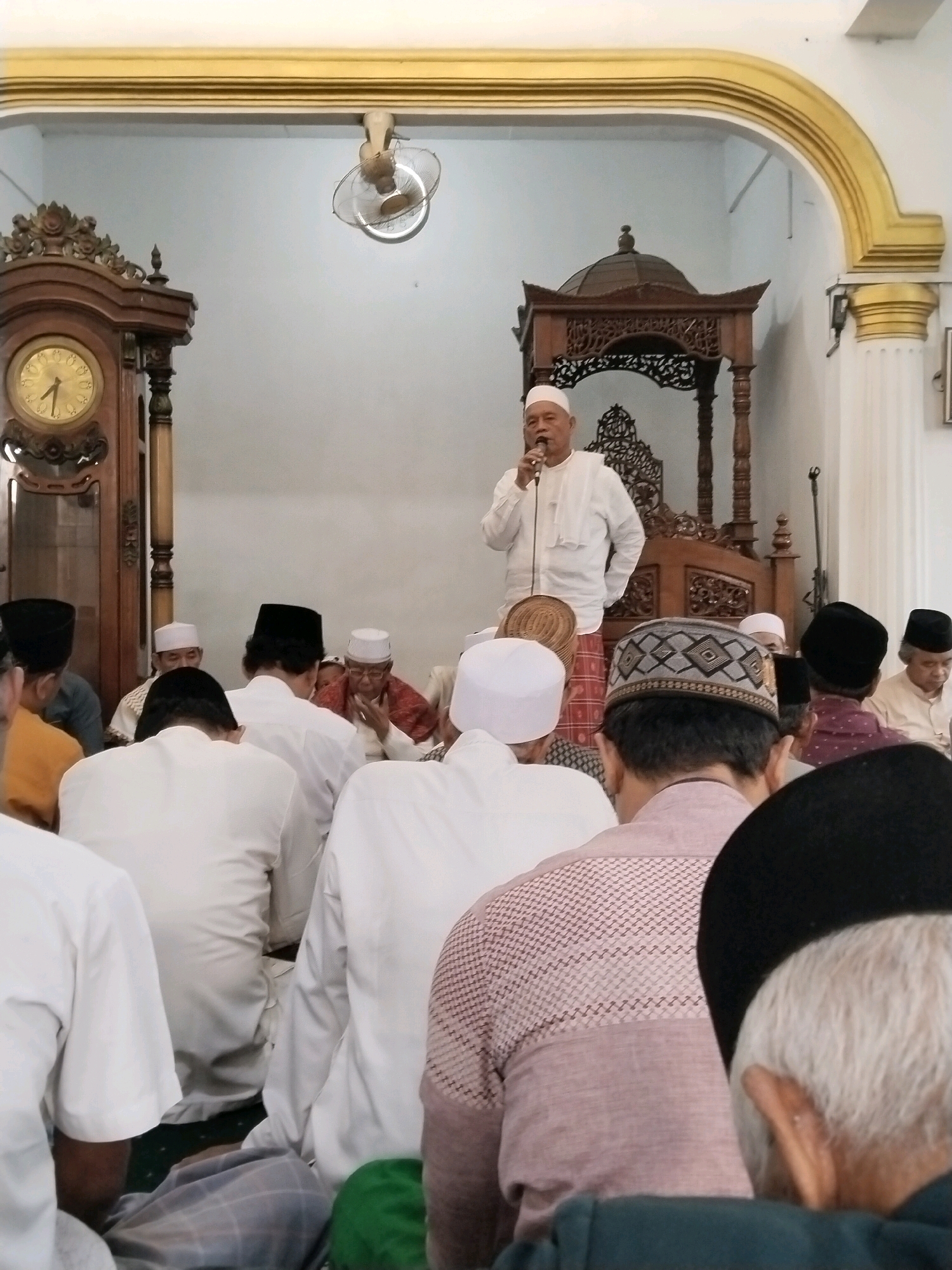 This image depicts an indoor gathering, likely a religious service, taking place in a room with a high, ornate ceiling and light-colored walls. A man wearing a white cap and white attire is standing at a wooden podium, speaking into a microphone. He appears to be the central figure addressing the attendees.
In the foreground and midground, a group of men are seated, facing the speaker. Most of them are wearing white caps or traditional Islamic headwear, and many are dressed in white or light-colored clothing. The men are varied in age, with some appearing to be elders. Their postures suggest attentiveness to the speaker.
Behind the speaker, a large, dark wooden floor clock stands to the left, its face visible. To the right of the speaker, a portion of a wooden structure, possibly a decorative screen or part of the room's architecture, is visible. A ceiling fan is mounted on the upper wall, suggesting the room might be warm.
The scene conveys a sense of community and religious observance. The environment appears to be a formal space, such as a mosque or a hall designated for religious gatherings. The specific location in the city of Serang, Indonesia, is not explicitly identifiable from the visual cues alone, but the attire and context are consistent with Islamic cultural practices common in Indonesia.