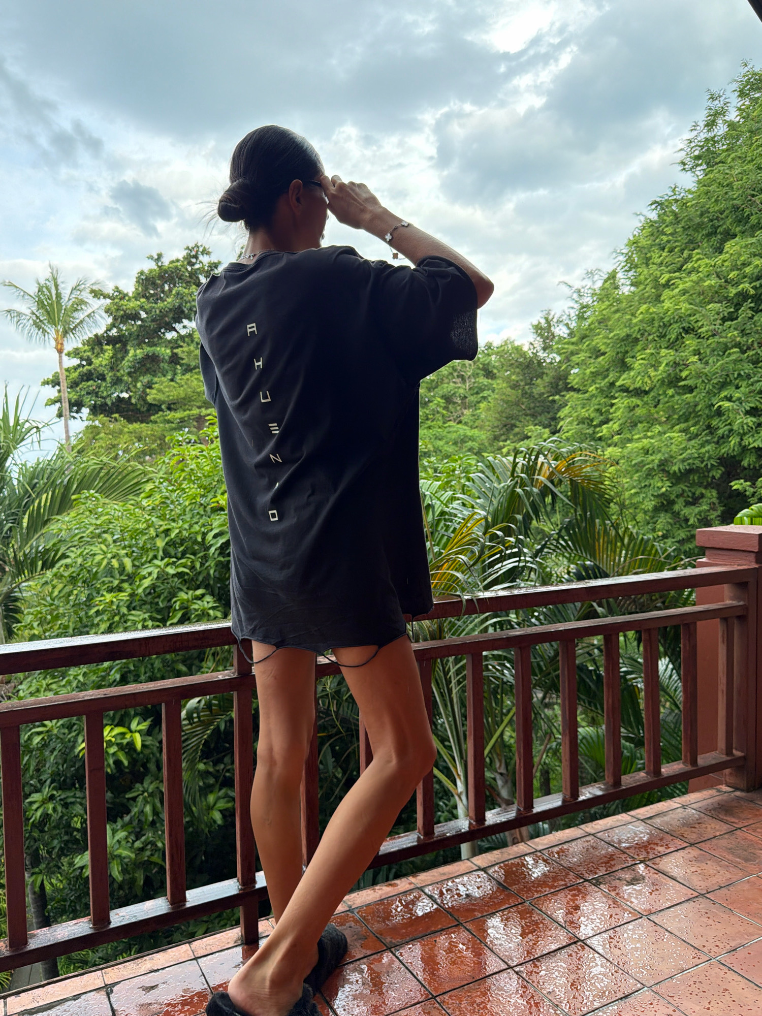A woman, viewed from behind, stands on a wet, tiled balcony overlooking a lush, green jungle. The sky is overcast with dramatic clouds. She wears an oversized black t-shirt with a white vertical graphic design on the back and dark, fuzzy slippers. Her hair is pulled back in a bun, and she is holding her hands up to her eyes as if looking through binoculars or shielding them from the sun. The balcony is furnished with a wooden railing, and the floor is covered in terracotta tiles that are wet and reflective, indicating recent rain. The background is filled with dense tropical foliage, including palm trees, suggesting a humid, possibly coastal environment in the Ko Samui District of Thailand.