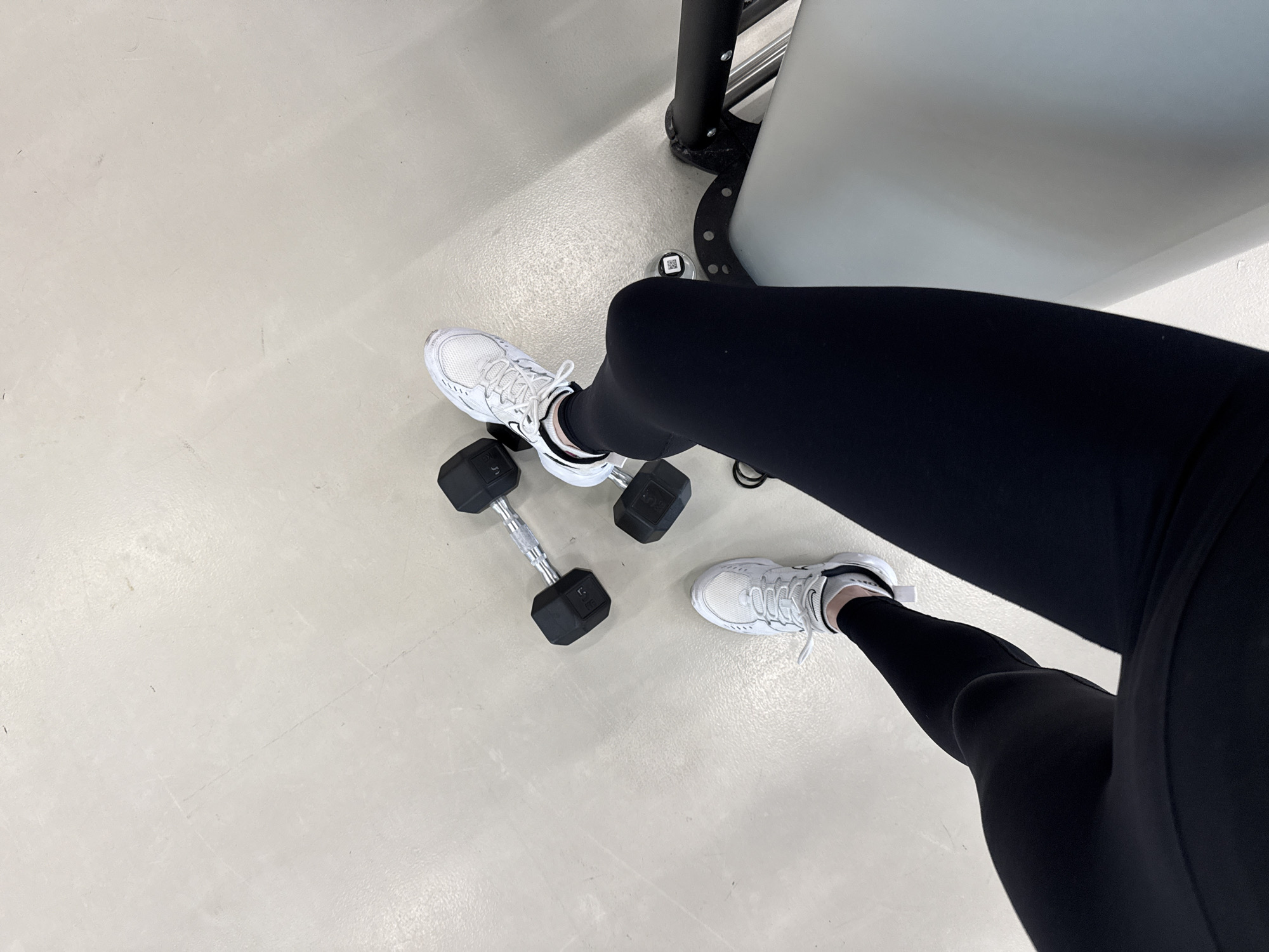 A high-angle, medium shot shows a person's legs in black leggings and white athletic shoes, with a pair of dumbbells nearby on a white floor, possibly in Unknown, Unknown. The dumbbells, with black ends and silver handles, are positioned to the left of the legs. The legs are slightly bent at the knees. Part of a gray workout machine is visible in the upper right corner of the frame.