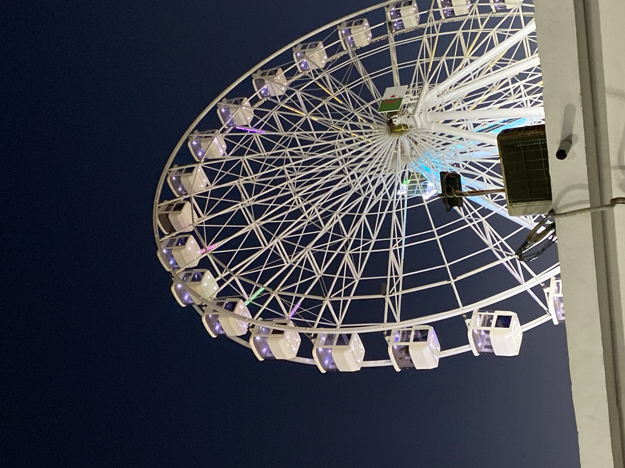 A low-angle, medium shot captures a large white Ferris wheel against a dark, clear night sky. The Ferris wheel is illuminated, with its cabins featuring glowing purple and pink lights. The structure of the wheel is intricate, with numerous spokes radiating from the center. At the hub of the wheel, an Algerian flag is visible. In the foreground, a white wall runs horizontally across the bottom of the frame. Partially obscuring the base of the Ferris wheel and resting on the wall are a black, rectangular object that appears to be an air conditioning unit, and a metal pole with a black square at the top. The overall scene suggests a night-time amusement park or fairground in the city of Hussein Dey, Algeria.