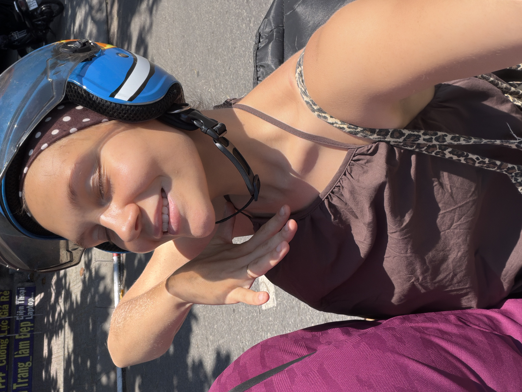 A woman wearing a blue helmet with white stripes and a brown polka dot headscarf is riding on the back of a motorcycle in Nha Trang Ward, Vietnam. She is smiling with her eyes closed and her left arm is raised, with her hand near her face. She is wearing a brown halter top and a leopard print strap is visible. To her left, a portion of a person wearing a purple garment is visible. The background shows a street with a sign in Vietnamese and shadows cast by trees.