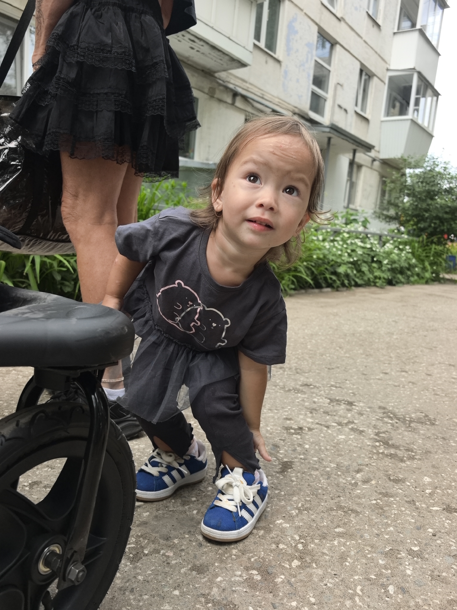 A young girl with light brown hair and brown eyes, wearing a dark grey t-shirt with two cartoon bears printed on it and dark grey leggings, is squatting near a black stroller in an urban outdoor setting. She is looking upwards and to the left with an inquisitive expression. The stroller's front wheel is visible in the foreground, partially obscuring the girl's legs. In the background, there is a light-colored, multi-story apartment building with numerous windows and balconies, and dense green foliage. The ground is paved with concrete. The presence of a stroller and apartment building suggests a residential area within the city of Chaikovsky, Russia.