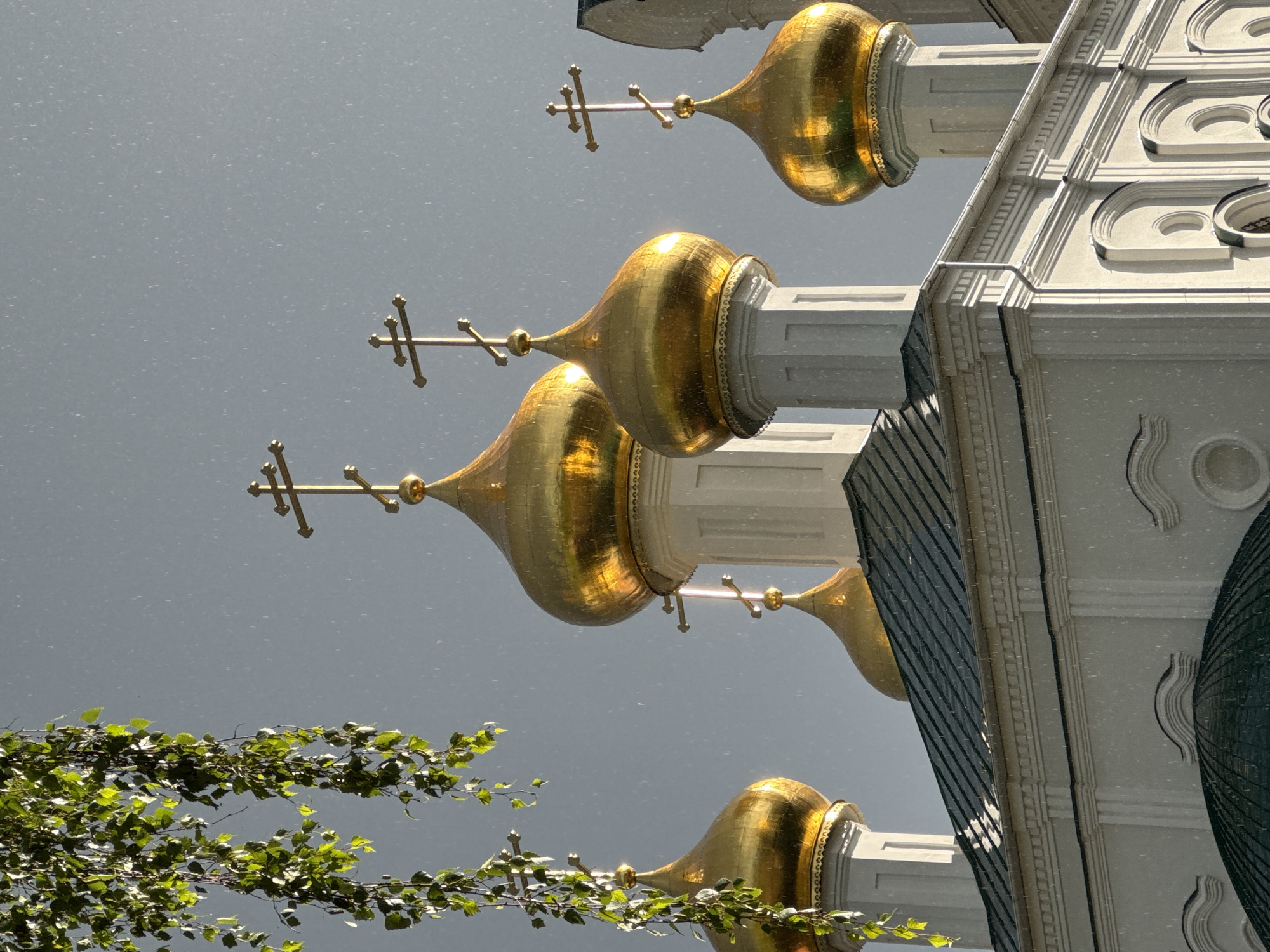 This media file showcases the ornate golden domes of a Russian Orthodox church, likely in Nizhny Novgorod, Russia. The perspective is unconventional, appearing rotated or reflected, with the sky as a backdrop. The church's architectural details are prominent, featuring white stone structures adorned with intricate moldings and decorative elements. The golden domes themselves are lustrous, reflecting the ambient light. Each dome is topped with a cross, a common symbol in Orthodox Christianity.
In the foreground, branches with vibrant green leaves suggest the presence of trees or foliage nearby, hinting at an outdoor setting, possibly a park or garden adjacent to the church. The weather appears overcast, with a grey, muted sky that allows the golden domes to stand out. Small white specks are visible throughout the image, which could be rain, snow, or dust particles, adding a textural element to the scene. The lighting is soft, indicating it might be daytime, but not necessarily direct sunlight. No people are visible, and the scene conveys a sense of architectural grandeur and religious solemnity. The composition is abstract due to the rotation or reflection, focusing on the interplay of light, shape, and texture. There is no visible text in the image.