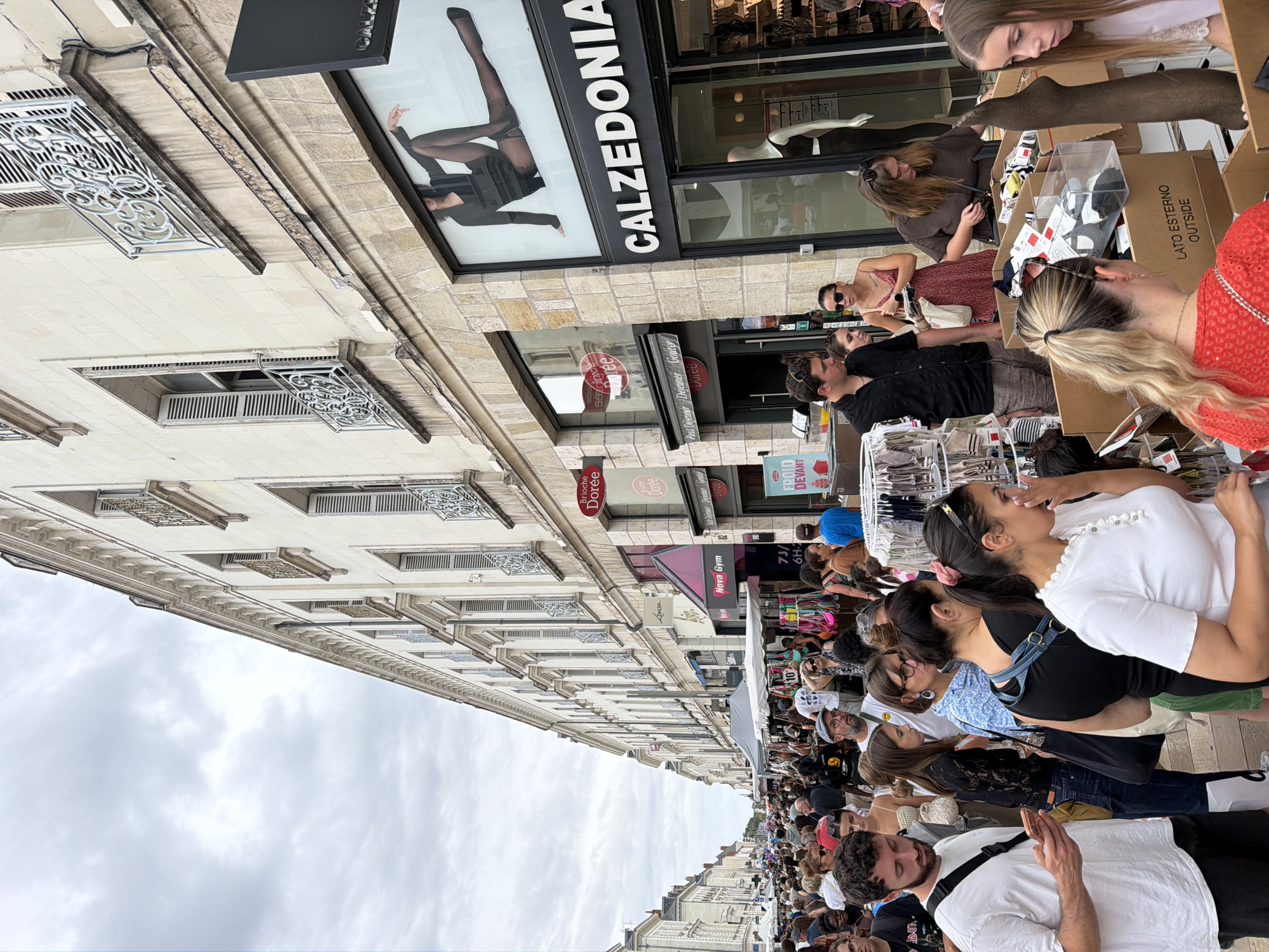 The image depicts a bustling street scene in Tours, France, likely during a market or festival. The foreground features a crowd of people browsing through various goods displayed in cardboard boxes and on racks. Among the merchandise are clothing items, possibly including socks or hosiery, with one display featuring a mannequin leg wearing dark stockings. Several individuals are visible examining the products.
The middle ground shows more stalls lining the street, with awnings and tents providing shade. The architecture of the buildings lining the street is visible, characterized by cream-colored facades, shuttered windows, and ornate wrought-iron balconies. Signage for businesses such as "Calzedonia," "Nova Gym," and "Brioche Dorée" are present.
The background extends down the street, revealing a long procession of buildings and a dense gathering of people continuing further into the distance. The sky is overcast with clouds. The overall impression is of a lively urban event with a significant amount of pedestrian traffic and commercial activity.