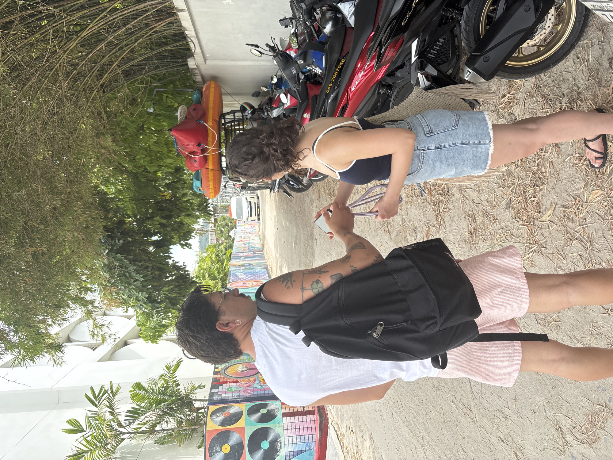 In Baan Chaweng, Thailand, a man wearing a white t-shirt and pink shorts, with a black backpack, stands facing away from the camera and interacting with a woman in a dark blue tank top and denim shorts. The woman is holding a mobile phone and a purple strap. They are standing on a sandy path with dried leaves. To their right, a row of parked motorcycles is visible. In the background, a building with a distinctive facade and a vibrant mural depicting vinyl records and colorful designs are present. Overhead, bamboo trees create a canopy. A vehicle with red and orange inflatable rafts attached to its roof is also visible in the distance.