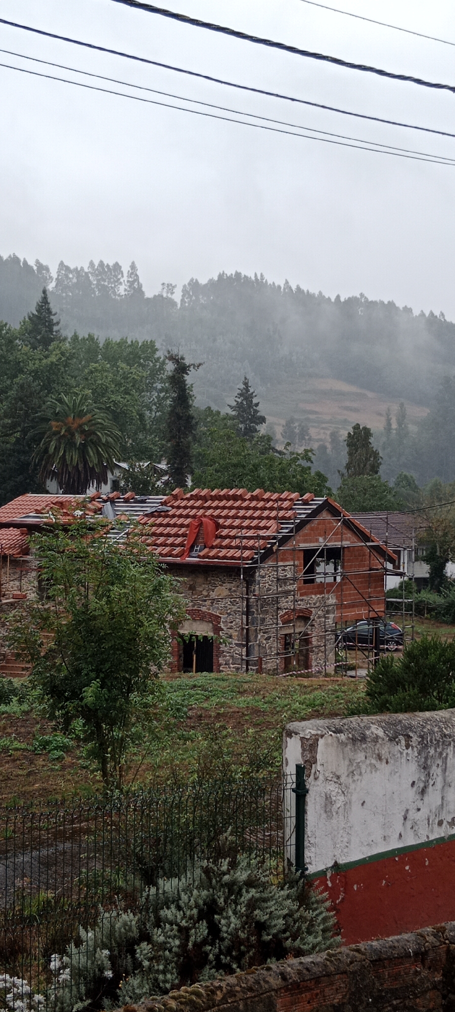 The image shows an overcast day with a misty mountain range in the background. In the mid-ground, a stone and brick building is under renovation, with scaffolding erected around it and the roof being retiled with orange tiles. A dark-colored car is parked to the right of the building. In the foreground, there is a wire fence and lush green vegetation. The overall scene depicts a rural or semi-rural setting with ongoing construction work. The location is the city of Pravia, Spain.
