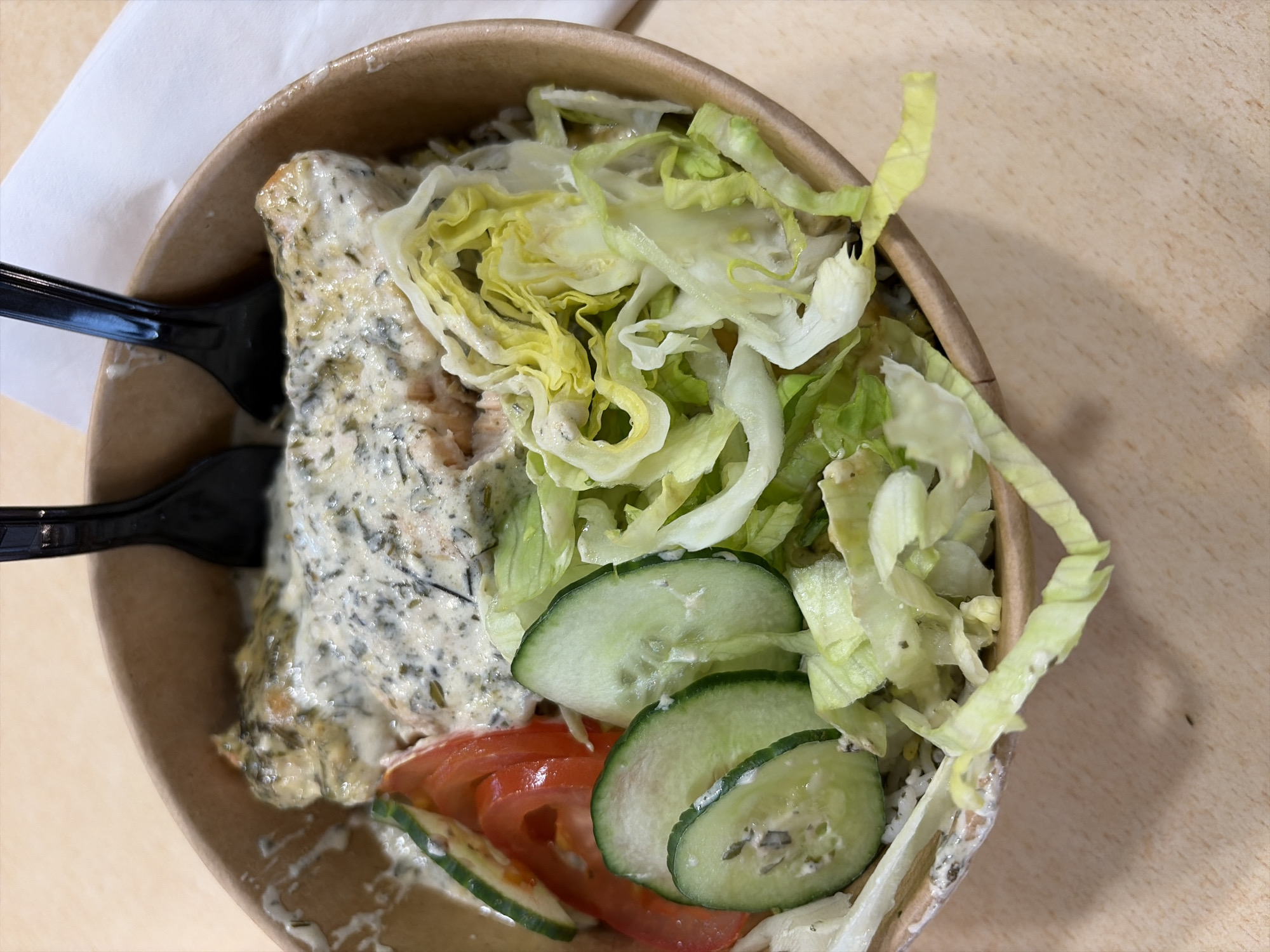 The image shows a takeaway container on a light brown surface, likely a table. The container is filled with food. The visible contents include a portion of salmon covered in a creamy sauce with green herbs. There is also lettuce, sliced cucumber, and sliced tomatoes. Two black plastic forks are resting in the container next to the salmon. A white napkin is partially visible near the container. The image gives no information about the location or city.