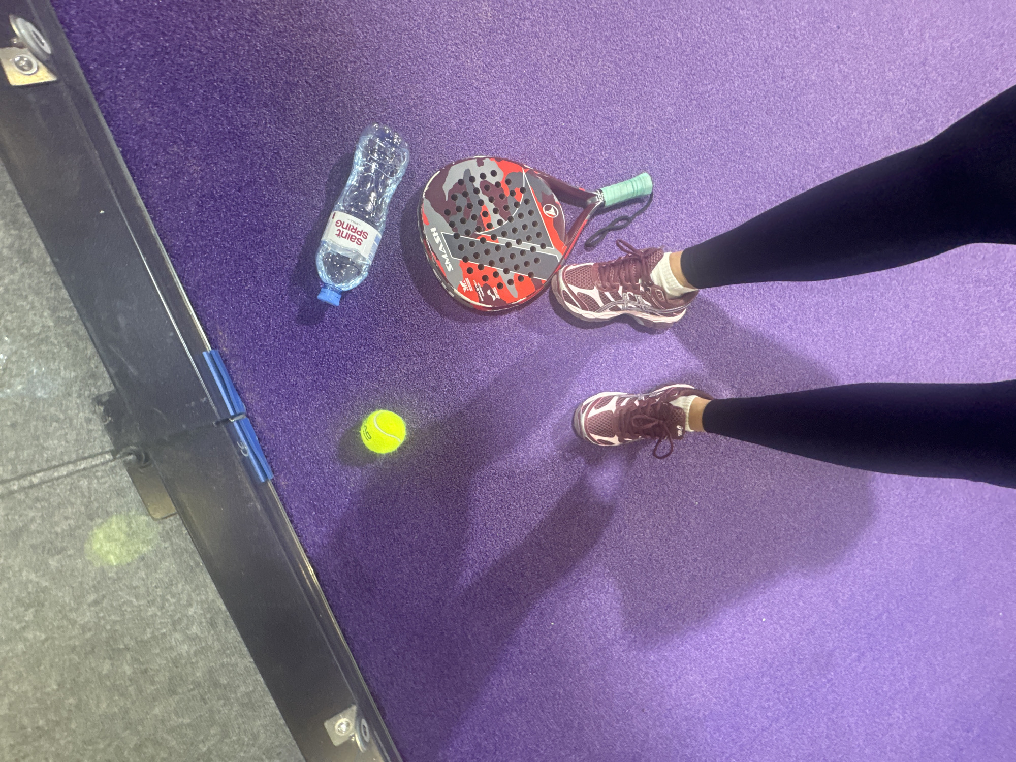 This overhead shot captures a person's lower legs and feet standing on a vibrant purple artificial turf court, indicative of a padel court. To the left, a dark grey textured surface meets the purple court, separated by a black structure that appears to be part of the court's boundary or enclosure.
On the court, a red, grey, and black padel racket with a camouflage pattern and a green grip is visible. The word "SMASH" is clearly printed on the racket's face. Next to the racket lies a clear plastic water bottle with a blue cap, featuring a label that reads "Saint Spring" (albeit upside down). A bright yellow padel or tennis ball, marked with "HEAD," rests near the racket and bottle.
The person is wearing dark leggings, white athletic socks, and maroon or dark red athletic shoes with white detailing, possibly from the Asics brand, though the logo is not fully legible. The lighting appears consistent, suggesting an indoor setting or a uniformly lit outdoor area without direct sunlight. The arrangement of the equipment and the person's stance imply a break during or after a game. The setting is in Krasnoyarsk, Russia.