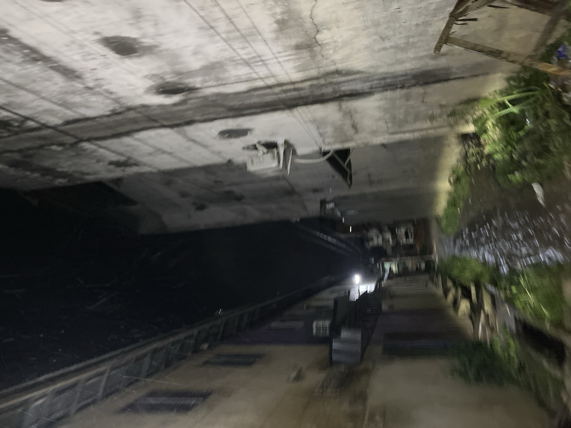 This image depicts an outdoor scene at night, likely in an urban or industrial setting, possibly near water. The perspective is from a height, looking down at an angle.
The main subject is a dark, possibly water-filled channel or ravine that runs diagonally through the frame. On the left side of this channel, there is a metal railing or barrier, and beyond that, a dark, flat expanse. On the right side of the channel, there are several elevated structures that appear to be part of a building or walkway. These structures are dimly lit, with some featuring windows or openings. Lush green vegetation is visible growing along the right edge of the scene, appearing somewhat wild or overgrown.
In the upper portion of the image, a concrete structure with visible rebar and some cracks suggests construction or a partially completed building. There are some utility boxes or fixtures attached to this structure.
The lighting is dim and artificial, with some areas illuminated by what appear to be outdoor lights, casting a glow on the structures and the vegetation. The overall mood is somewhat somber and perhaps abandoned, due to the darkness and the raw, unfinished appearance of some elements. There is no visible text. The location context provided places this scene in Kota Kuala Simpang, Indonesia. The time of day is clearly night.