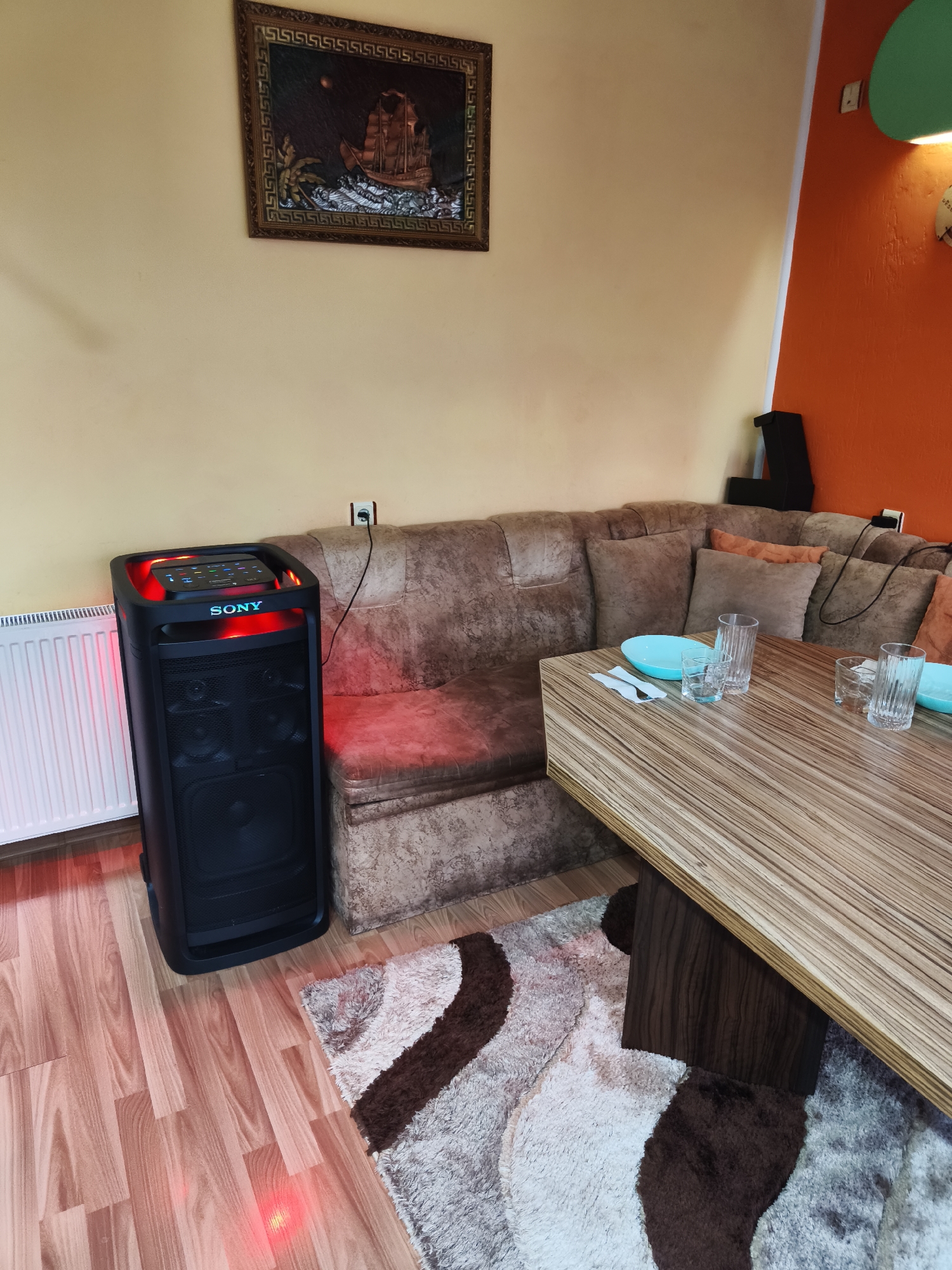 The image depicts an indoor scene, likely a dining or living area. A large, black Sony speaker with red and blue lighting is prominently featured on the left side of the frame, positioned next to a white radiator. The speaker is on a light-colored wooden floor. Behind the speaker and to the right is a large, brown sectional couch with numerous throw pillows. A wooden dining table with a light-colored, striped wood grain is in the foreground and extends to the right, partially covered by a shaggy rug with brown and cream patterns. On the table are several drinking glasses, two light blue plates, and silverware. In the background, a framed artwork of a sailing ship hangs on a beige wall. To the far right, an orange wall is visible. The scene appears to be an interior domestic setting.