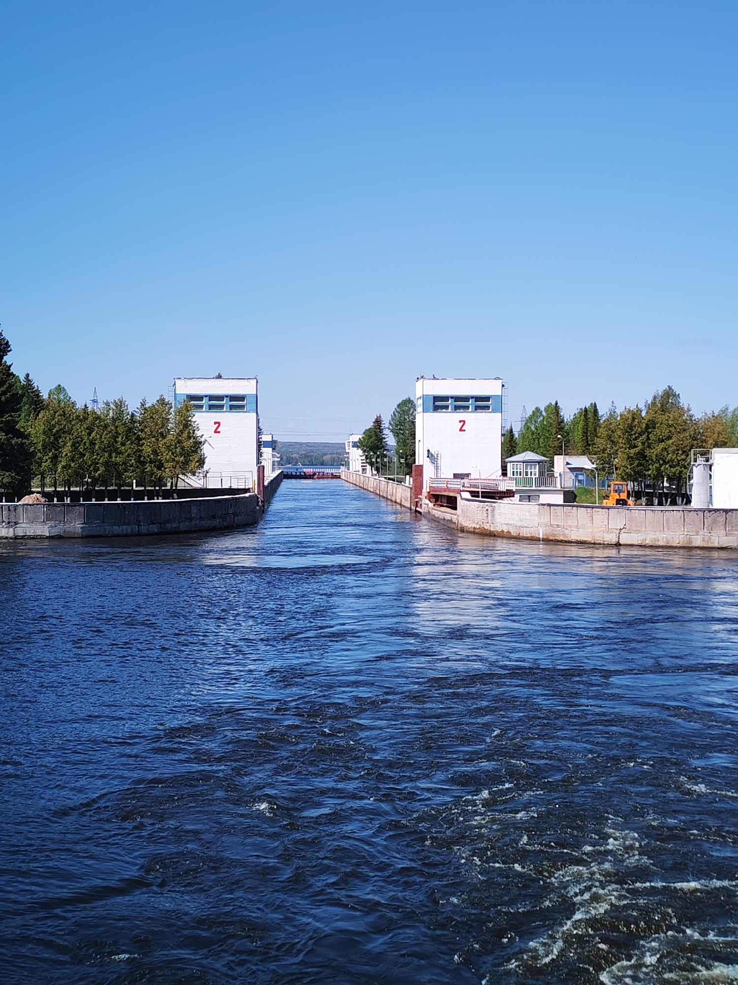 The image shows a canal lock in Белоусово, Russia. Two white buildings with the number "2" in red stand on either side of the canal. Concrete retaining walls line the canal banks. The water in the canal is dark blue and slightly turbulent. Trees are visible on both sides of the canal. The sky is clear and bright blue.