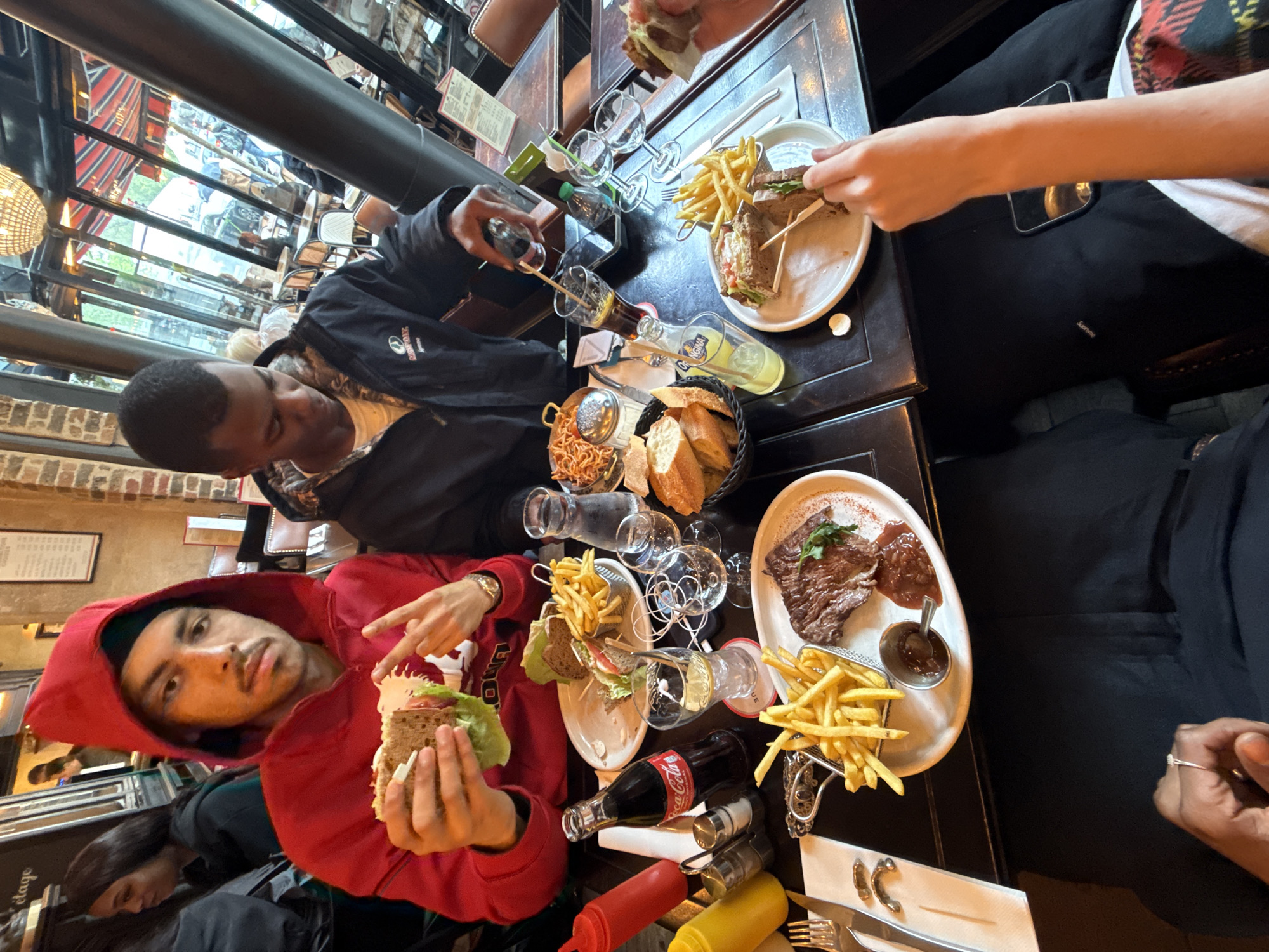 The image captures a moment at a restaurant in Paris, France, likely near a window given the street view and bright daylight streaming in. Two individuals are prominently featured at the table. One person wears a red hooded sweatshirt and holds a sandwich, pointing towards it, with another person seated next to them. Adjacent to them, a second person is pouring a beverage from a glass bottle.
The table is laden with food. There are plates of sandwiches and French fries. The table also contains bottles of Coca-Cola and other beverages, various sauces, salt and pepper shakers, and glassware. A plate with steak and French fries, as well as a basket of bread, sits to the right.