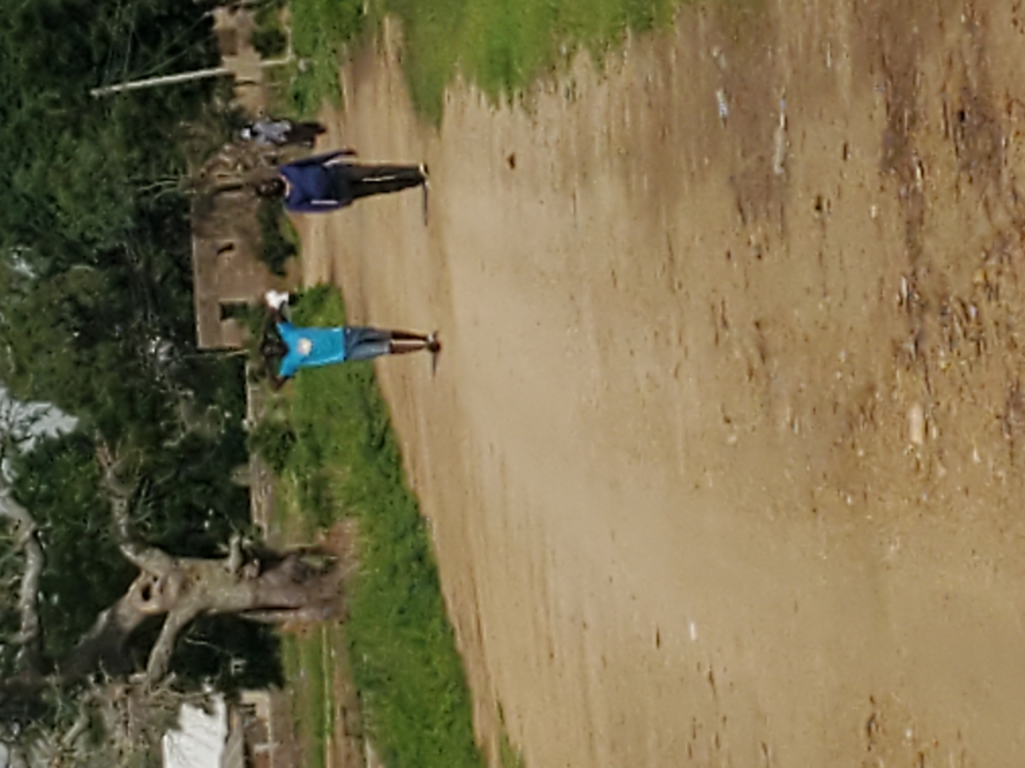 In a dirt road, two men are walking away from the camera. The man on the left is wearing a blue t-shirt and shorts. He has his hands on his head. The man on the right is wearing a blue long-sleeved shirt and dark pants. In the background, there are trees and buildings. The man on the left is standing in front of the man on the right. A third person in a dark shirt is walking in the distance.