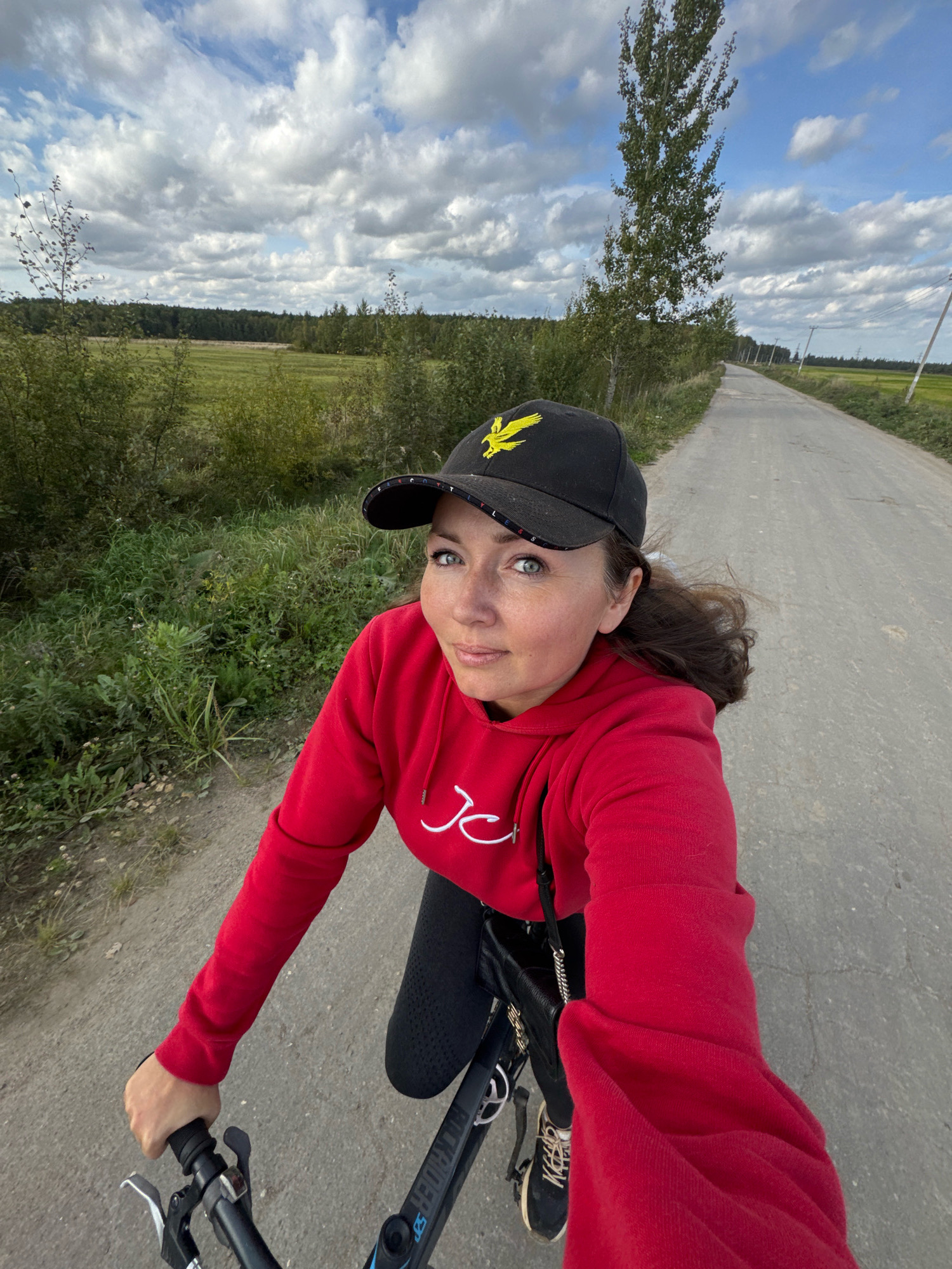 A woman wearing a red hoodie and a black baseball cap with a yellow eagle logo is riding a bicycle down a paved road. The road is lined with grass and trees. In the background, there is a field and a blue sky with white clouds. The woman is looking at the camera and smiling. The location is the city of Innolovo, Russia.