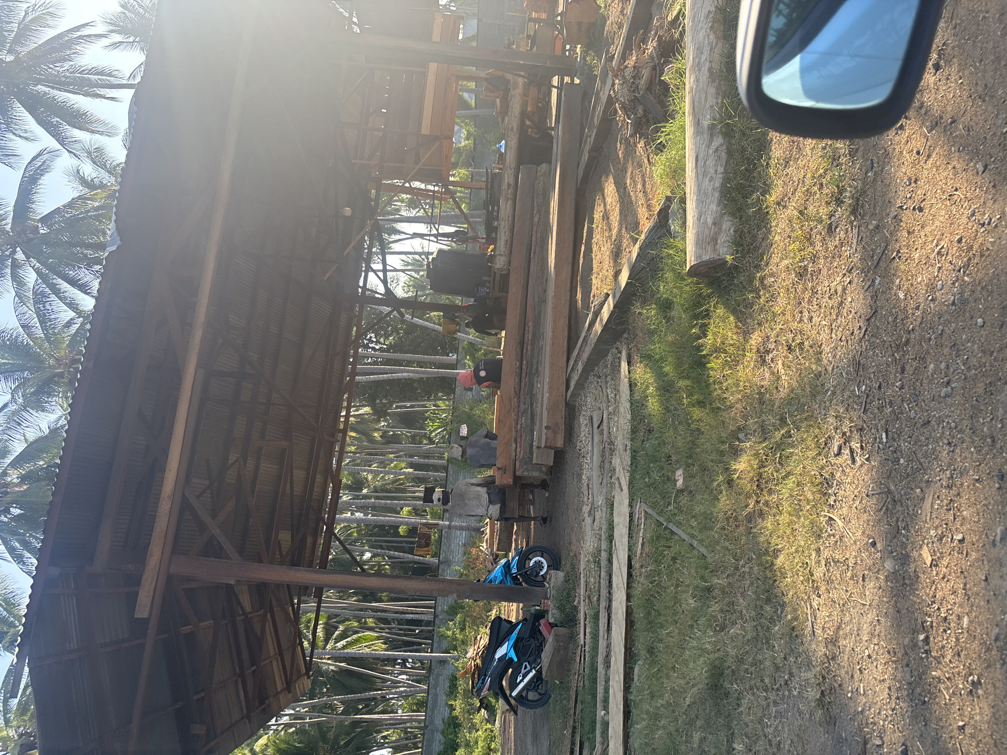 The image depicts a sawmill located in Bunto, Indonesia, under a covered structure with a corrugated metal roof supported by wooden beams. Several people are present, some appearing to be workers. In the foreground, a blue and black motorcycle is parked on the left. The ground is a mix of dirt and grass, with numerous large wooden logs and planks scattered throughout the scene. In the background, a dense arrangement of palm trees is visible against a clear sky. The scene is illuminated by natural sunlight, which creates strong shadows and lens flare, particularly on the right side of the image.