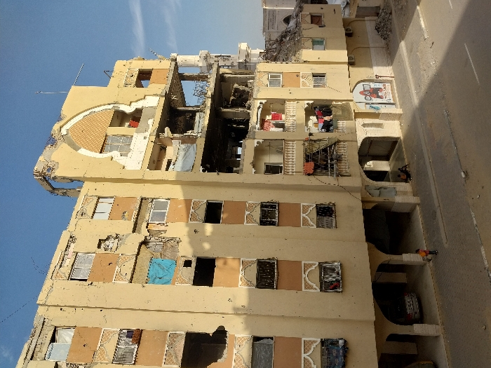 The image displays a severely damaged multi-story residential building in Khan Yunis, Palestinian Territory, under a clear blue sky. The light beige structure exhibits extensive destruction, including large holes penetrating walls, shattered windows, exposed interiors, missing balcony sections, and a partially collapsed roofline. Numerous smaller pockmarks and potential bullet holes cover the facade. A prominent arched architectural feature on the upper right side is significantly compromised. Laundry hangs on several balconies, notably on the right side of the building, and a blue tarp covers a window opening on the third floor from the bottom on the left.
At ground level, the building features arched open spaces. A silver car is parked in one such area on the left. A child, wearing an orange top, stands on the paved sidewalk directly in front of the building. To the right, two individuals are seated on the ground within a shaded arched recess. Debris is scattered on the street and along the base of the building, where red graffiti is also visible on a wall section.