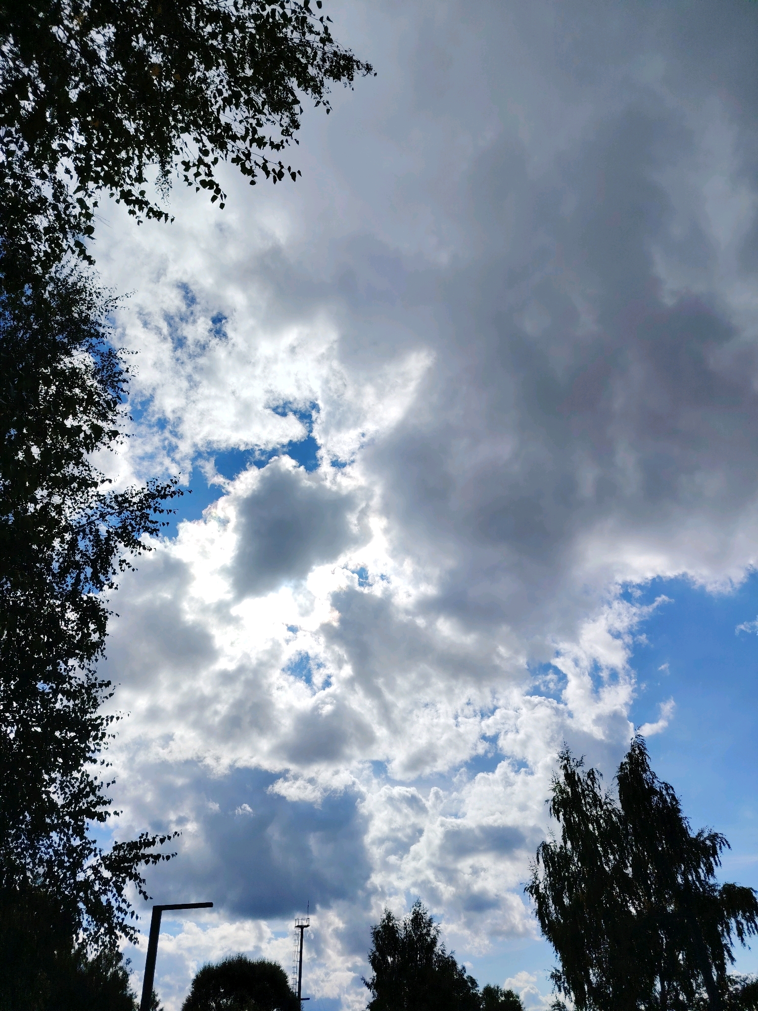 The image shows a cloudy sky over the city of Konakovo, Russia. In the foreground, silhouetted trees frame the view. To the left, a lamppost is visible. Further to the right, a tall structure resembling a communication tower stands against the sky. The clouds are a mix of dark gray and white, with patches of blue sky peeking through. The overall impression is of an overcast day with intermittent sunshine.