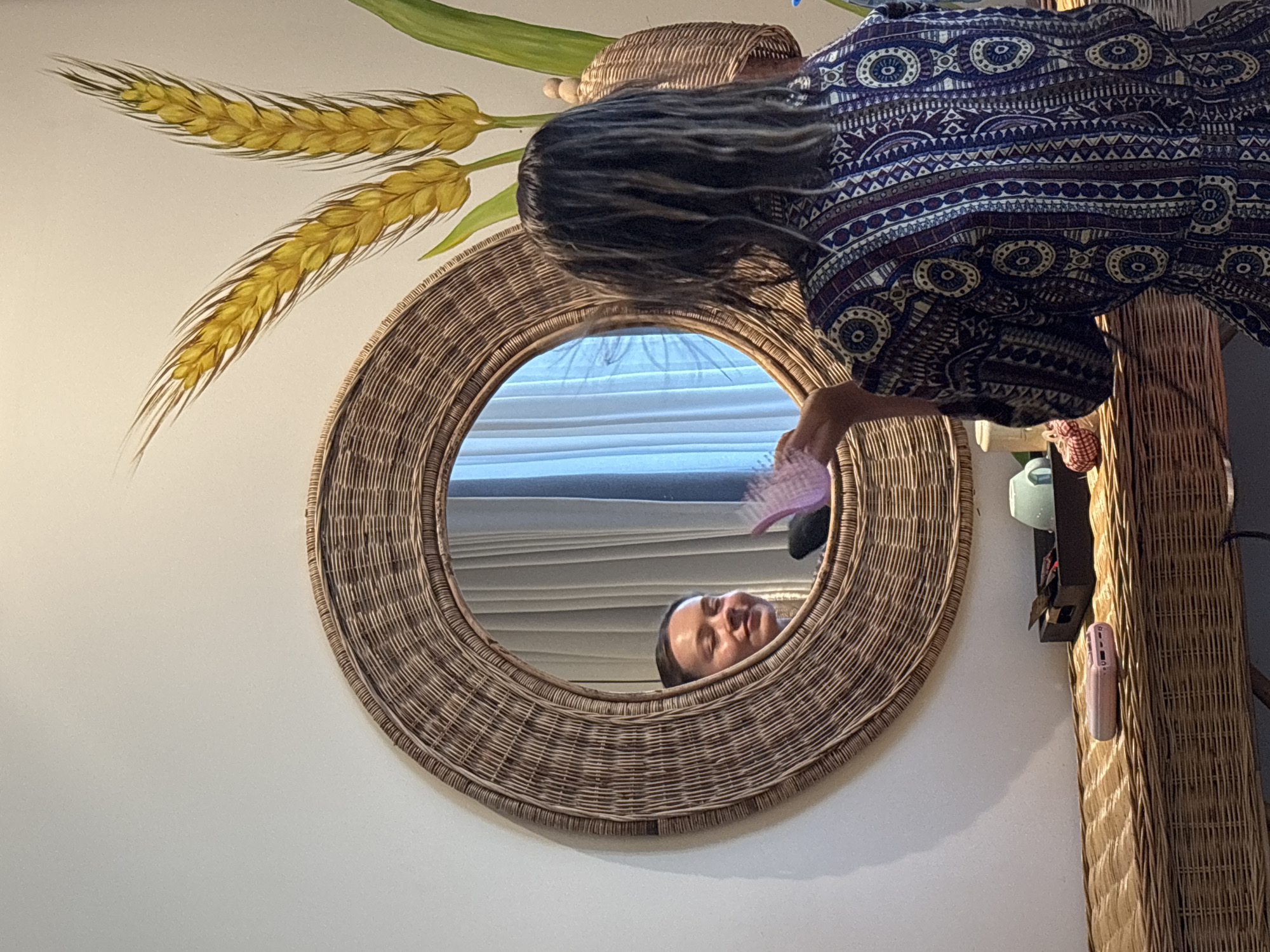 A woman brushes her hair in the mirror. The mirror is oval shaped, woven, and hanging on a wall. A mural of wheat is painted next to it. The setting is Nha Trang Ward, Vietnam.