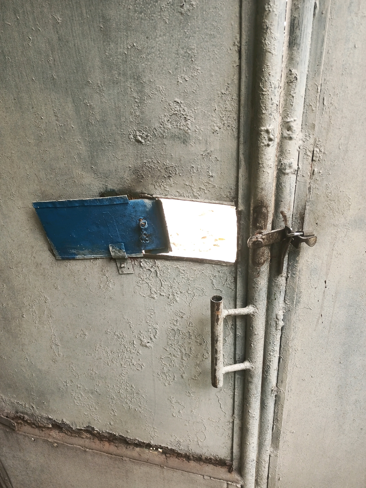 The image shows a close-up of a weathered, gray metal door with peeling paint. A small, blue flap is open on the left side of the door, revealing a bright, possibly sunlit opening behind it. A metal latch, appearing rusted, is visible on the right side of the door, secured to a vertical pipe or bar. A section of a U-shaped metal handle is also visible on the left side of the door. The door appears to be part of an exterior structure. The image does not provide any contextual clues to determine its specific location in the city of Malang, Indonesia.