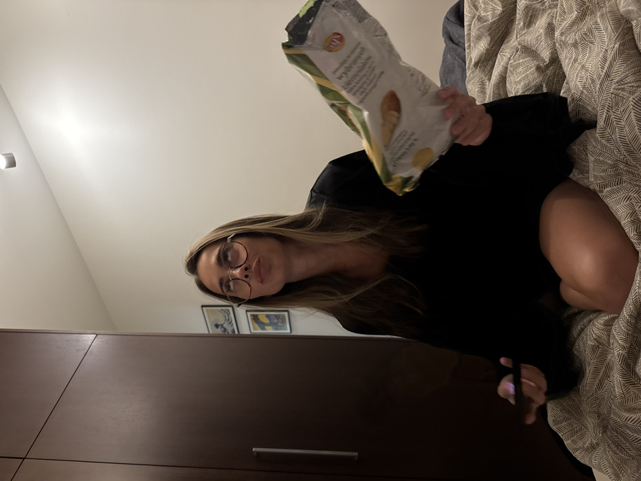 A woman with long blonde hair and glasses sits on a bed with patterned sheets. She wears a black long-sleeved shirt and holds an open bag of Lay's potato chips. A dark wood wardrobe stands to her left. Three framed pictures hang on the wall behind her. The room is lit by a light fixture in the upper left corner.