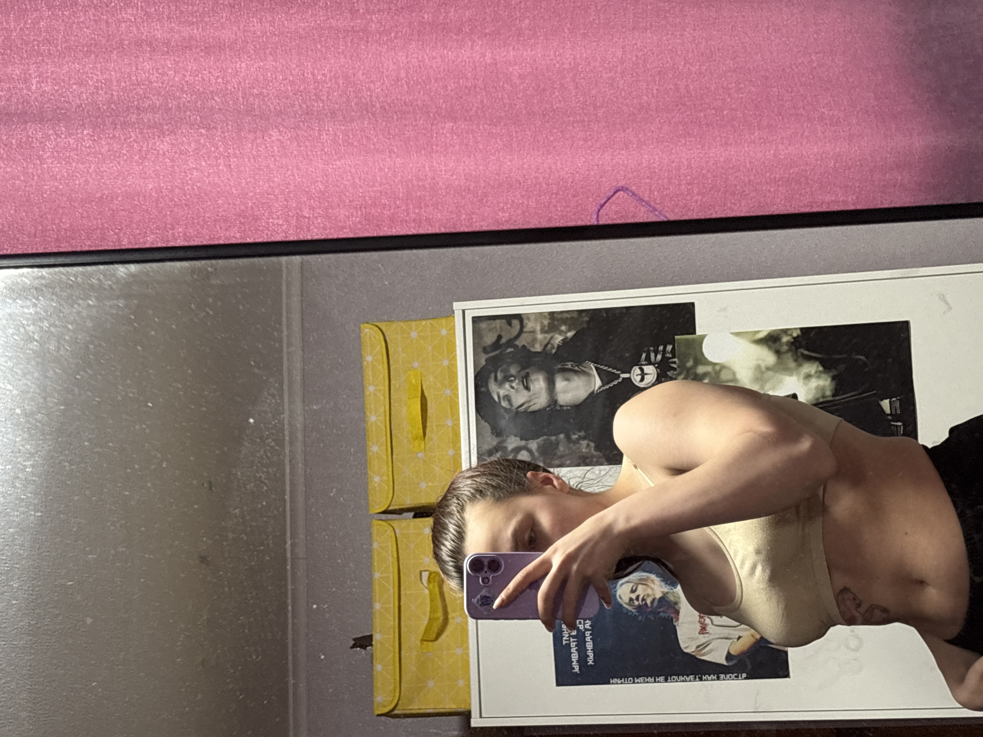 The image depicts a woman taking a mirror selfie. She is holding a purple smartphone, with its screen showing an image of a person, partially obscuring her face. She is wearing a beige tank top and dark shorts. In the mirror's reflection, behind her, are yellow storage boxes with a banana pattern and a framed black-and-white photograph of a person. The top of the image shows a pink surface, possibly a shelf or headboard. The background includes a plain, light-colored wall. The location is stated as Прикамский, Russia, but no element in the image confirms this location.