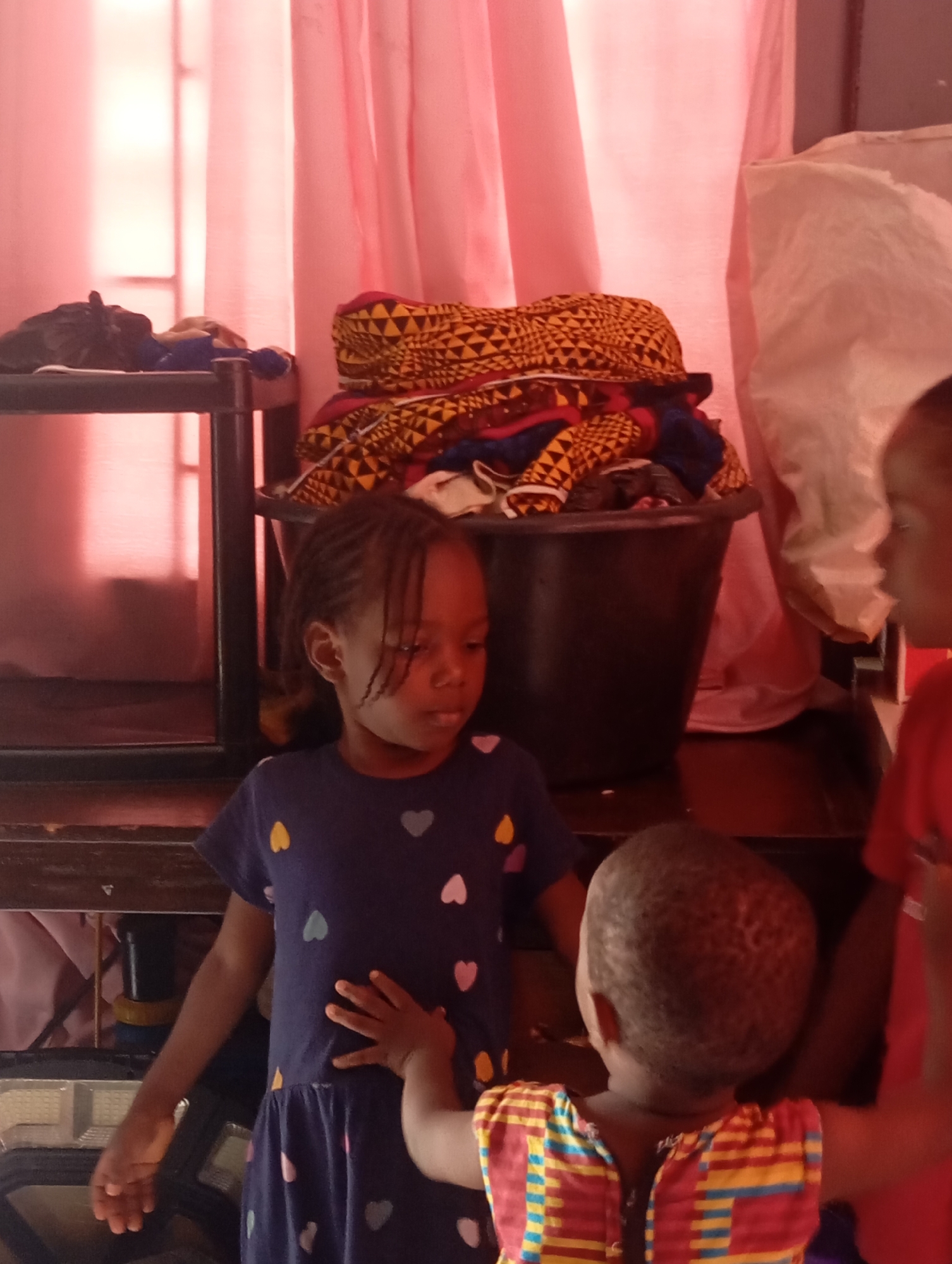 In Nsugpe, Nigeria, two young children, likely sisters, are interacting in a room with pink curtains. The older child, facing the camera, wears a dark blue dress adorned with multi-colored heart shapes and has her hair braided. Her right hand rests on her stomach. Beside her, a younger child, seen from the back, wears a vibrantly patterned shirt with horizontal stripes in red, yellow, and blue. In the background, a black bucket is filled with colorful fabric, possibly intended for sewing or crafts. To the left, a dark shelf unit is visible, and to the right, a large, light-colored plastic bag sits. The lighting suggests the image was taken indoors during the daytime.