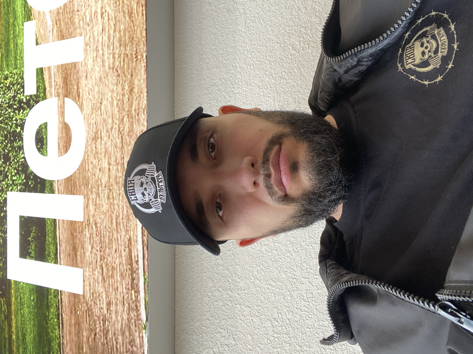 This portrait-style image, rotated 90 degrees clockwise from its upright orientation, features a man looking directly at the camera. He has a dark beard and mustache and maintains a neutral, calm expression.
The man is wearing a black baseball cap with a white logo on the front, depicting a skull, barbells, and the word "BARBARIAN". He is also dressed in a black zippered jacket over a dark top, which features a gold-colored logo on the left chest. This logo is similar in design to the one on his cap, showing a skull with barbells and the word "BARBARIAN" encircled by a barbed wire motif.
The background behind the man is split. To his left (when upright), there is a large sign or poster with a vibrant green, leafy pattern at the bottom and a brown, wood-like texture above. Large white letters are partially visible on this sign, including an "e" and an "o", which appear to be part of the word "Metro" or "Leto". To his right (when upright), there is a plain, light-colored wall with a textured finish, possibly stucco.
The lighting indicates it is daytime, but no specific weather conditions are discernible. The man appears to be posing for a photograph, with no other discernible activity. The location is Izhevsk, Russia.