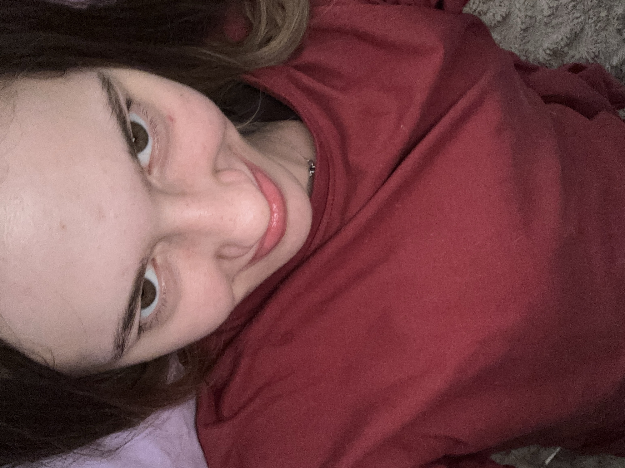 A person is lying on their back, wearing a red shirt. The camera is positioned above them, looking down. The person's face is visible, with their eyes looking upwards. Their lips are slightly parted, and they appear to be smiling. The background is a blurred mix of red fabric and a grey, textured surface. There is no discernible city or location information within the image.