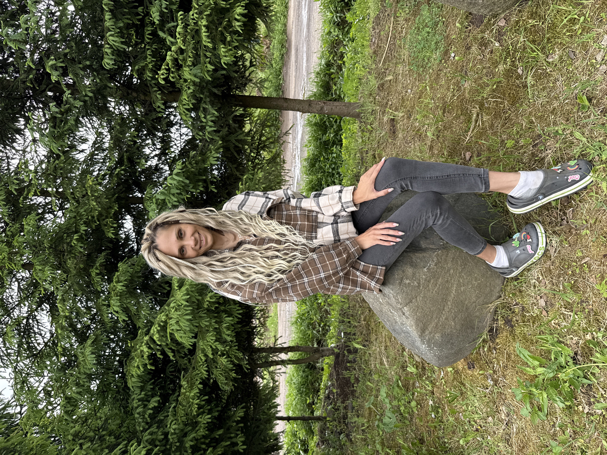 A woman is sitting on a rock in a grassy area with trees. She has long blonde hair and is wearing a plaid shirt, dark jeans, white socks, and grey shoes with embellishments. The area appears to be a park or natural setting. There is a dirt road and several small trees visible behind the woman. While Rusanovo, Russia is not discernible from the provided image, the scene could reasonably be located there.