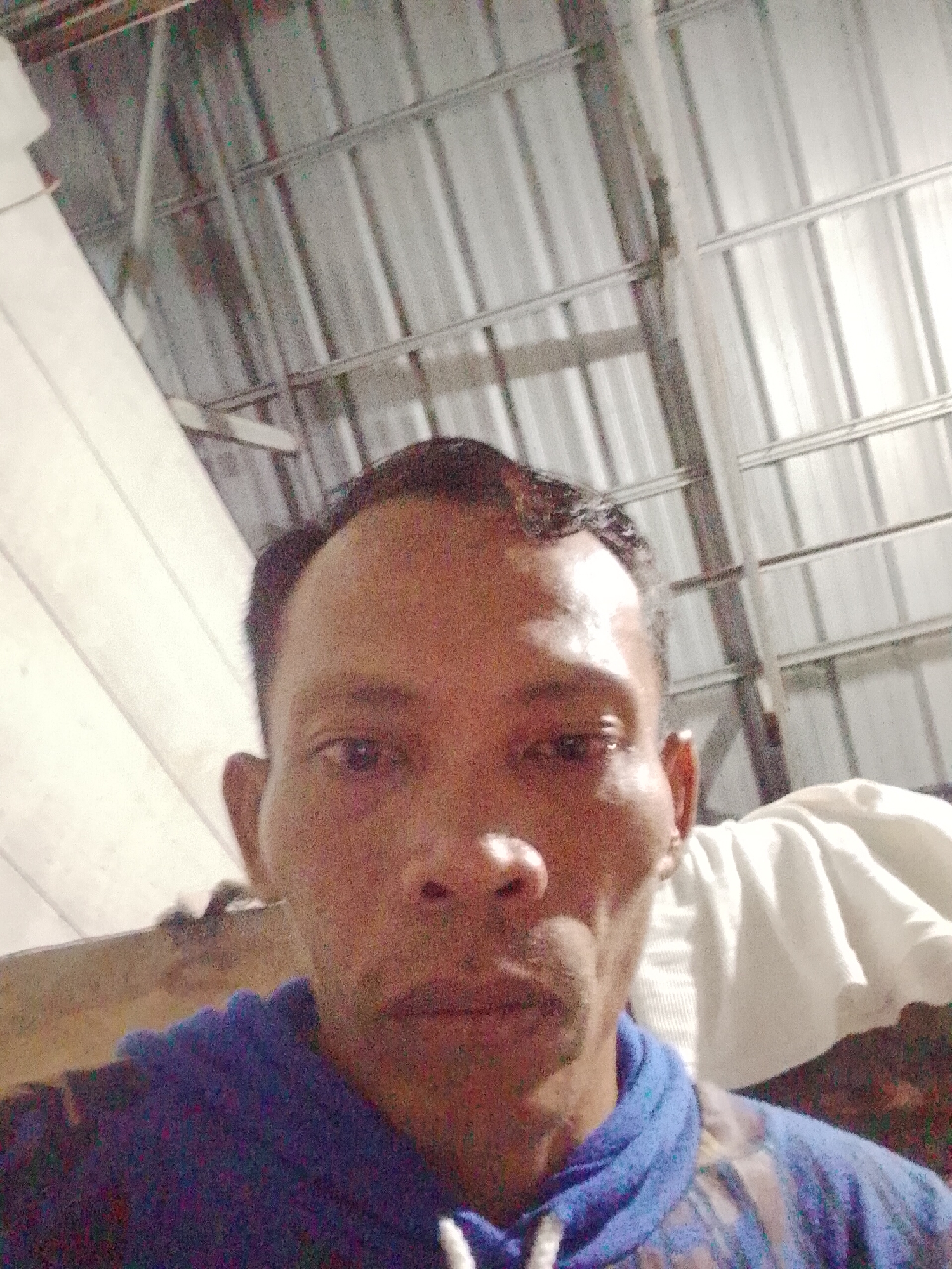 The photograph shows a man from a low angle, presumably a selfie. He has dark hair and skin, with visible facial features. He is wearing a blue hooded sweatshirt. The background is blurry, but appears to show the interior of a building or structure. The ceiling is made of metal and there is some sort of white cloth or material visible behind him. The photograph provides insufficient information to determine the specific location within Bukit Ketok, Indonesia.