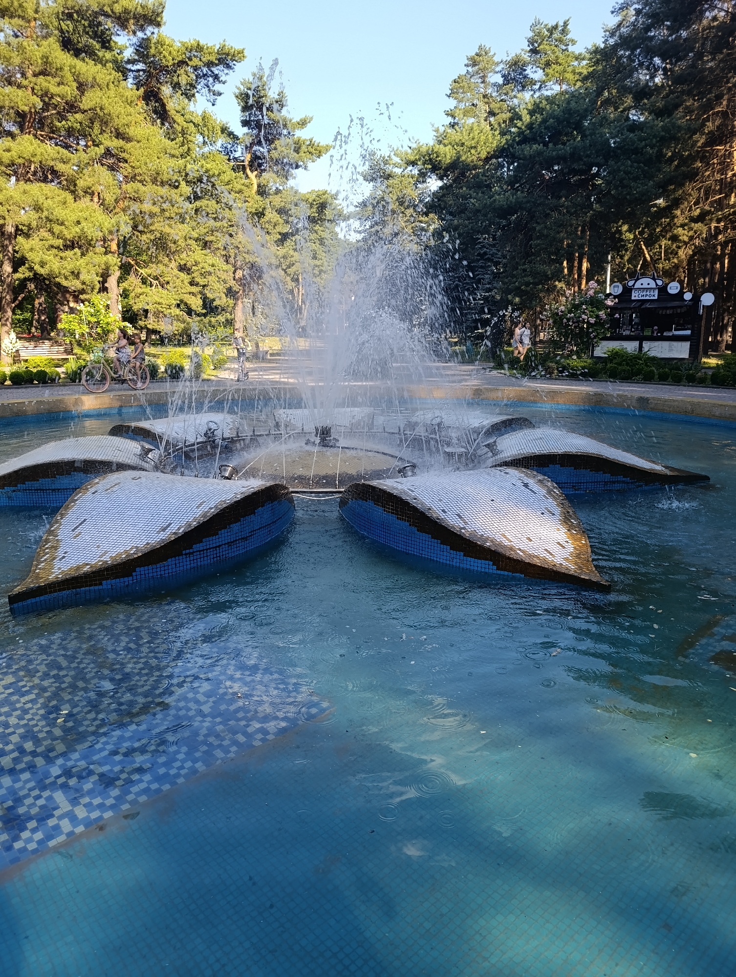 A fountain with flower-shaped petal elements, covered in small mosaic tiles, is active with water jets. The fountain is situated in a tiled pool with a checkerboard pattern of blue and white tiles. In the background, a park setting is visible with tall pine trees, a pathway, benches, and a small kiosk with a sign depicting a cartoon monkey. Two people are riding bicycles on the left side of the frame, and a person is standing near the kiosk on the right. The sky is clear and blue.