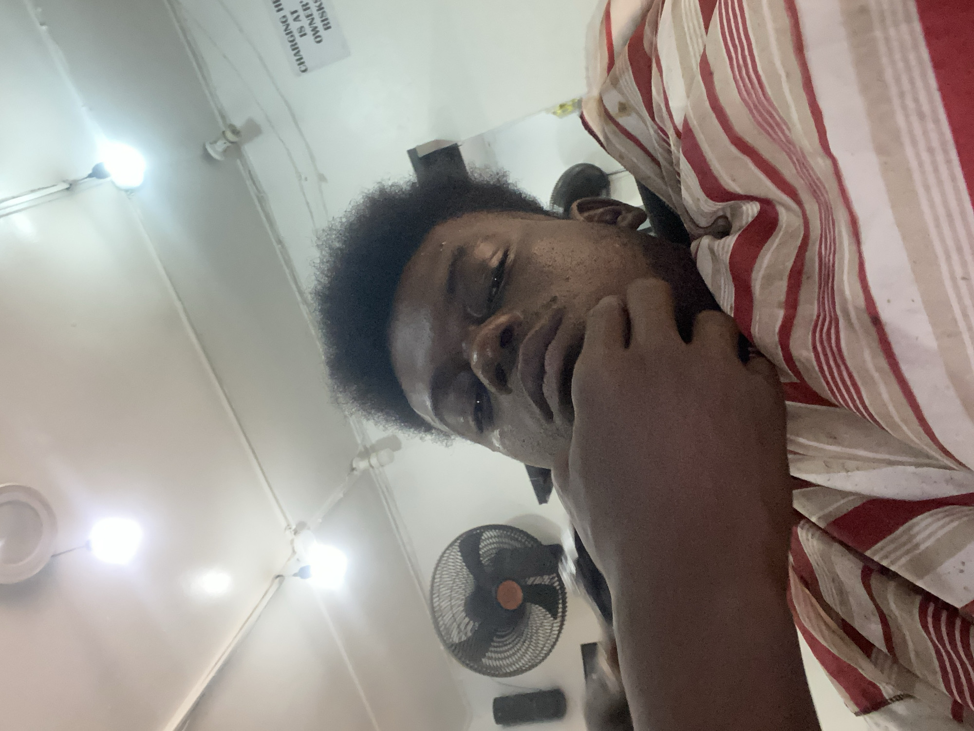An image, rotated ninety degrees counter-clockwise, depicts a person indoors, looking directly at the camera. The individual has dark skin and short, curly dark hair, and is wearing a striped shirt with alternating red, white/cream, and light brown vertical stripes. One hand is gently resting near their chin, giving a thoughtful or relaxed expression.
The setting appears to be a brightly lit indoor space with white or light-colored walls and ceiling. Several light sources are visible on the ceiling; some are glowing intensely, indicating bright illumination. A black wall-mounted fan is prominently featured in the background, below and to the right of the person's head. Below the fan, a small, dark rectangular object is mounted on the wall. In the upper left corner of the image, a white sign with black text is partially visible on the ceiling. The legible text on the sign reads: "CHARGING IS AT OWNER'S RISKS". The lighting conditions suggest it is likely daytime or a well-lit interior. The overall composition is a selfie or close-up shot taken from a low angle, emphasizing the person and parts of the room's interior.