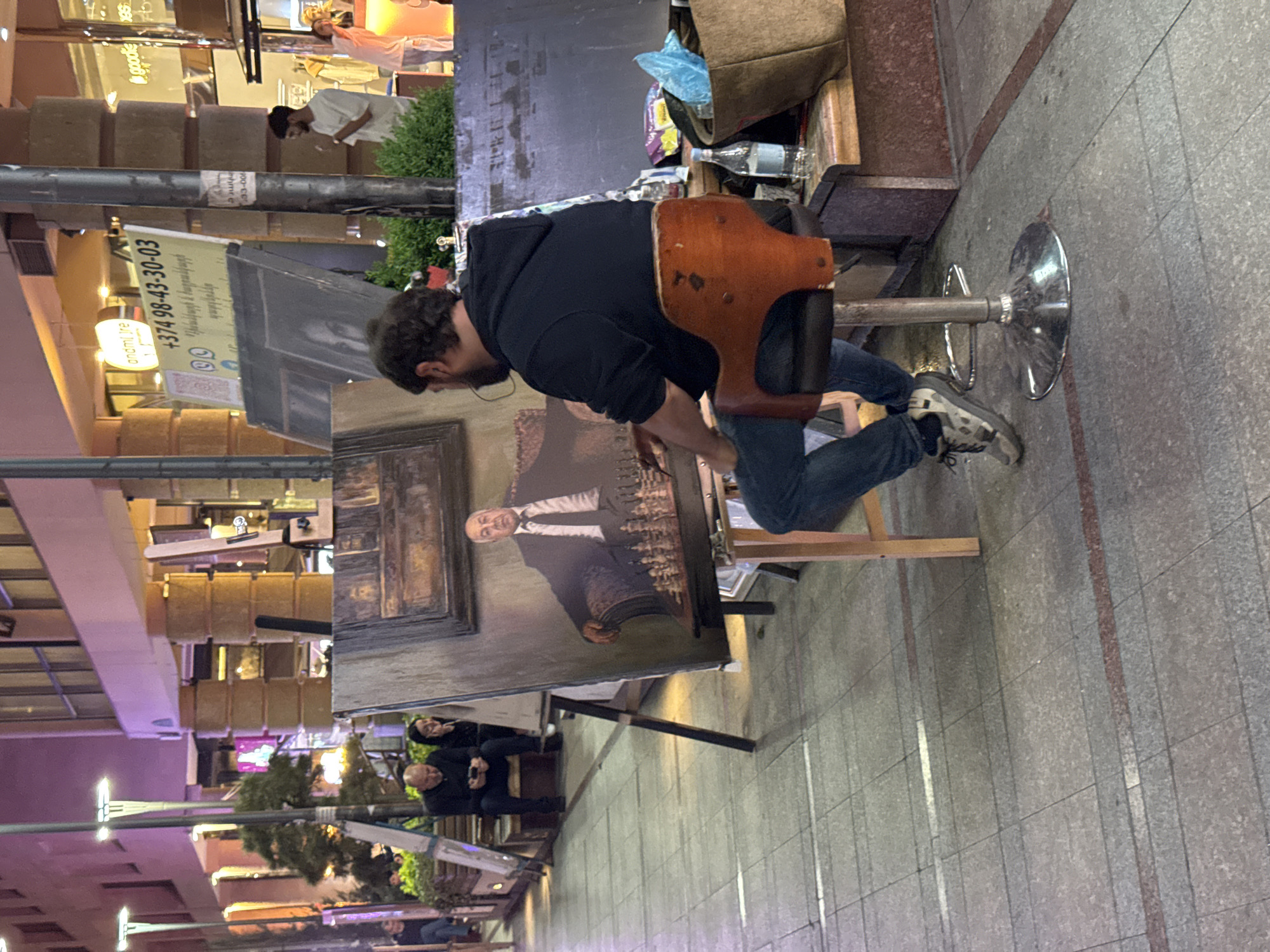 A street artist sits on a stool painting a portrait on an easel on a Yerevan, Armenia, sidewalk. The artist, a man with a dark beard and wearing a black hoodie and jeans, leans toward his work. The portrait appears to be of a man in a suit. Several people are sitting nearby. A light post with a sign displaying a phone number in Armenian is visible in the background. Buildings and a shop are visible in the background. The sidewalks are tiled.