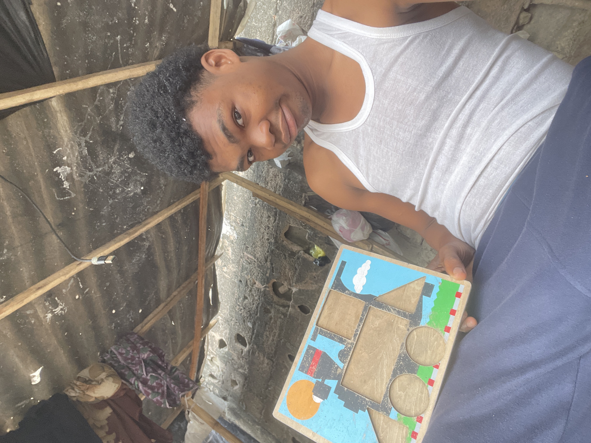 A young Black male, wearing a white tank top and dark blue pants, is sitting indoors and holding a wooden puzzle with a train design. The puzzle features a blue sky with clouds and a sun. The background shows a rough, textured wall and a corrugated metal roof supported by wooden beams. There are some cloth items and plastic bags visible in the background. The scene appears to be in a humble dwelling, possibly in the city of Ilado, Nigeria, characterized by its rustic construction and the presence of everyday items.