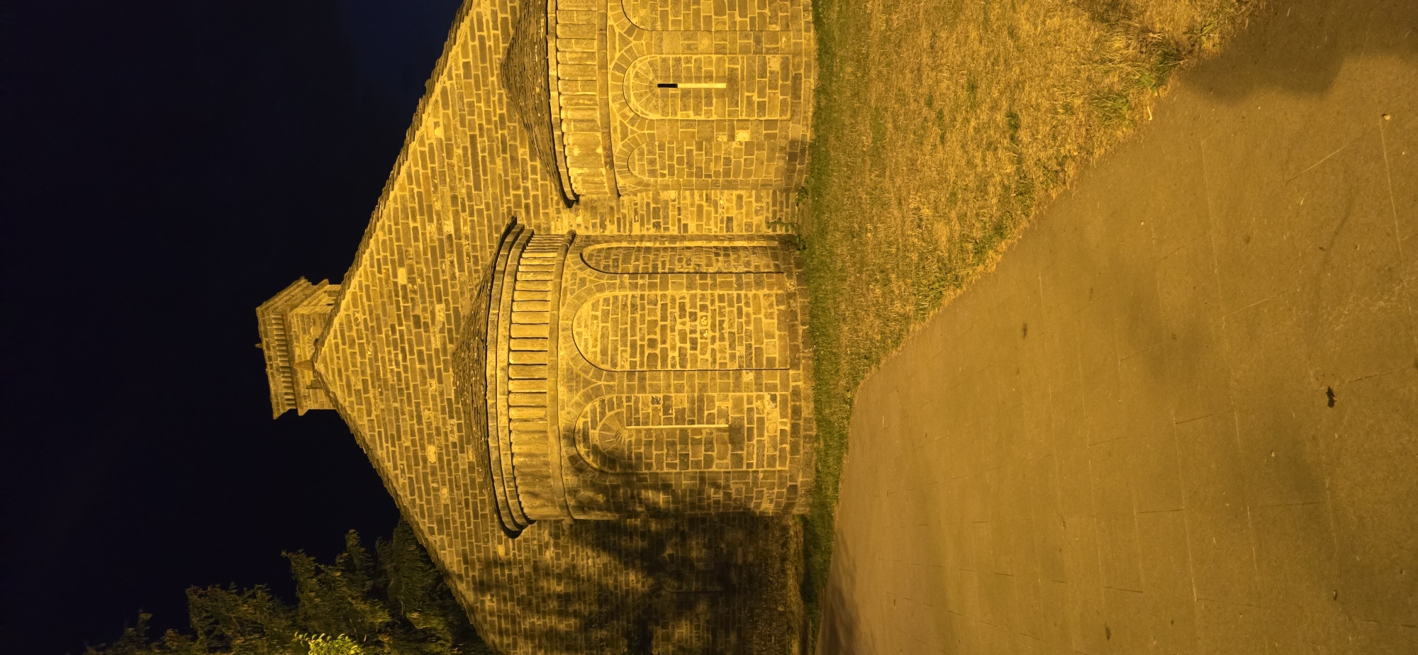 The image shows the exterior of a building constructed from yellow brick, illuminated by artificial light at night. The building has a steep, gabled roof and features two semi-circular apses on its facade, each with arched windows. A tall, rectangular structure, possibly a chimney or bell tower, is visible at the apex of the roof. To the left of the building, a portion of a pine tree is visible against the dark night sky. A paved walkway runs along the foreground, bordering a patch of grass. The overall scene is consistent with the Romanesque architecture found in Sallent de Gállego, Spain, a town known for its well-preserved churches and historical buildings.