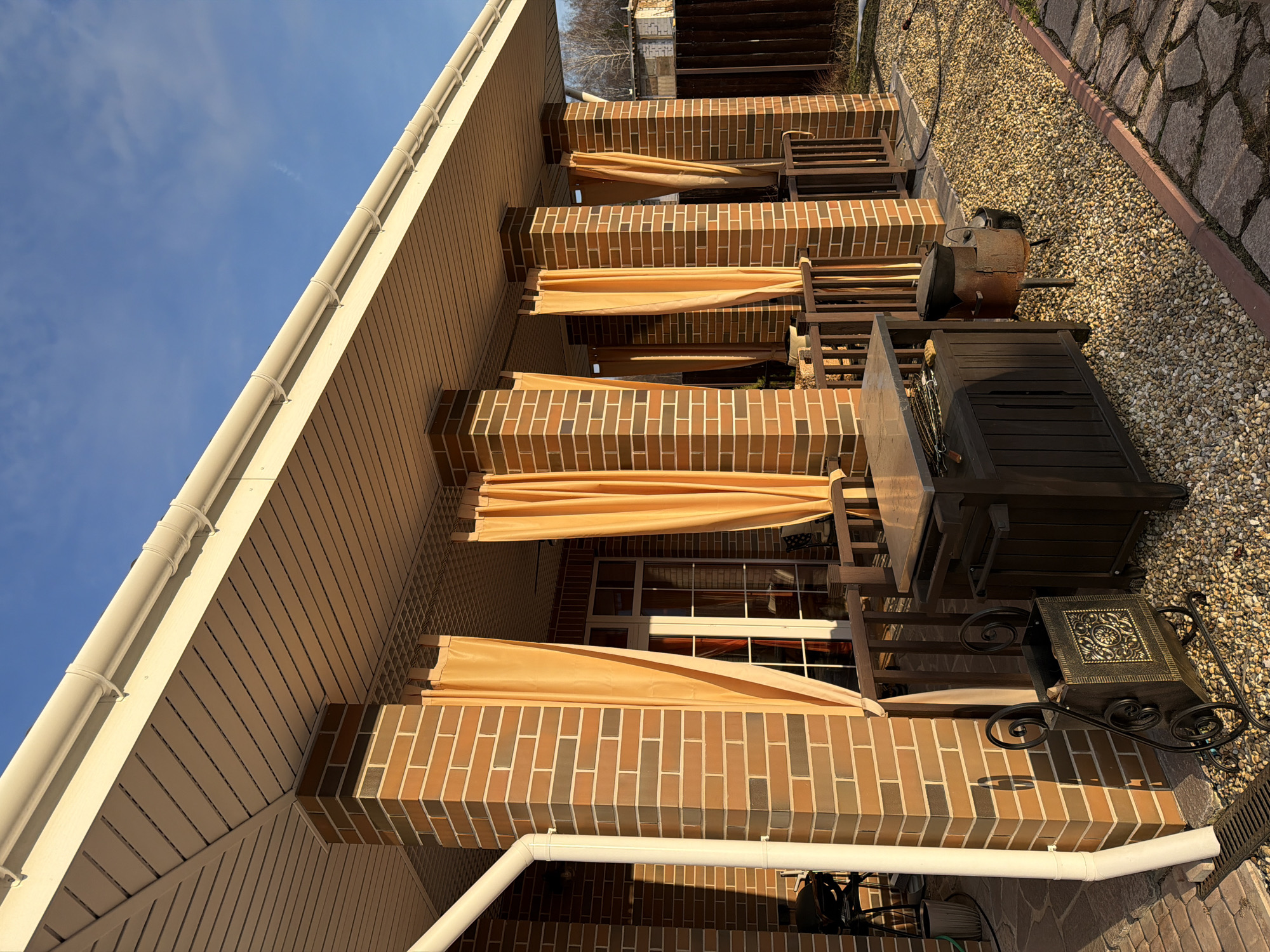 A low-angle shot shows the exterior of a brick building with a series of arched awnings. The awnings are a light yellow color and appear to be retracted, revealing the brickwork underneath. The brickwork is a mix of brown and tan shades, forming a rectangular pattern. A white rain gutter runs along the edge of the roof, which is angled toward the sky. The sky is a clear blue. To the right, outdoor furniture, including a table and a grill, is visible on a gravel surface, bordered by paving stones. The location is likely in Ryazan, Russia, based on the context provided.