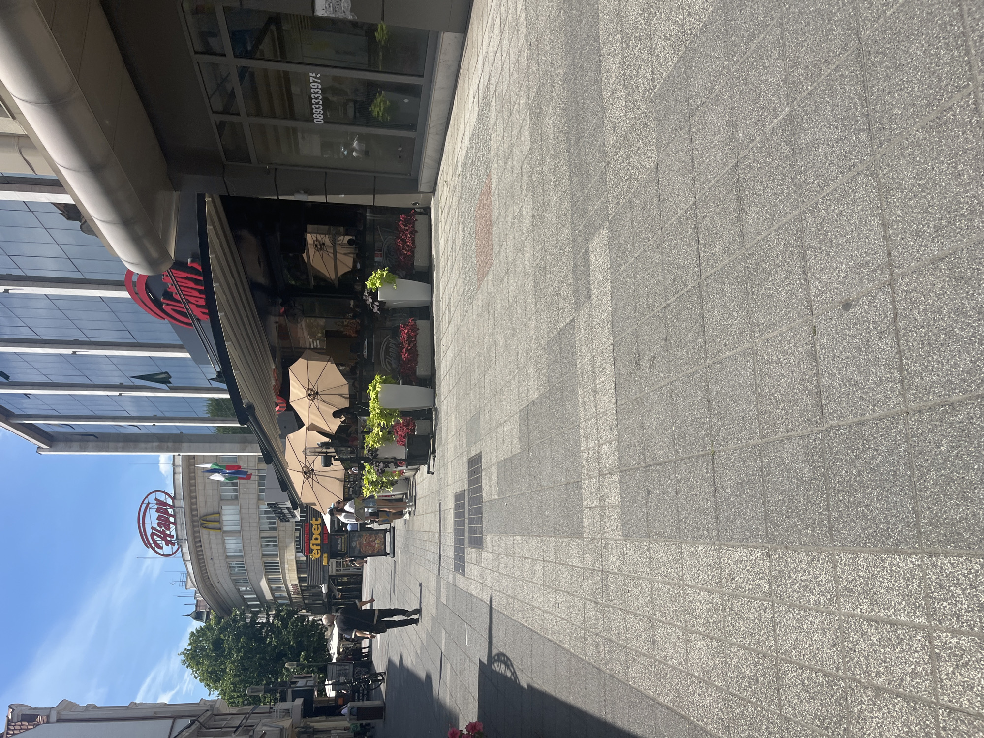 The image shows a street scene in Plovdiv, Bulgaria, specifically in the pedestrian zone near the "Happy" restaurant. In the foreground, a man in a black shirt and pants walks away from the camera on a tiled sidewalk. To the right, the "Happy" restaurant has an outdoor seating area with tables and umbrellas. In the background, a building with a large "Happy" logo is visible, as well as a McDonald's sign and Bulgarian flags. The sky is blue with some clouds. The scene appears to be a sunny day in a busy urban area.
