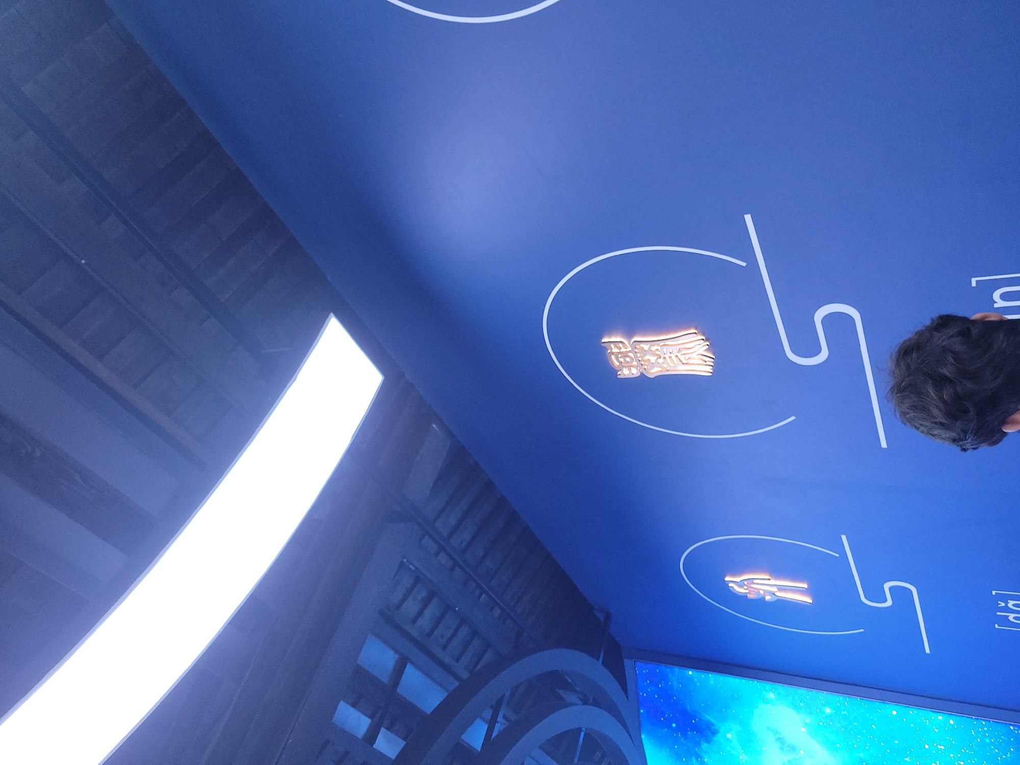 The image depicts a blue-toned interior setting, possibly a museum or exhibition space in Hechuan, China. The focus is on a blue ceiling adorned with white circular and angular line drawings, within which are embedded gold-toned emblems or symbols. A bright, elongated light source is visible on the left side of the frame, along with what appears to be a section of a building or display structure featuring repeating arch-like elements. A portion of a screen displaying a bright blue, cloud-like image is seen in the lower part of the picture. The top right corner shows a glimpse of a person's dark hair. The scene suggests a modern, possibly artistic or informational environment, likely indoors.